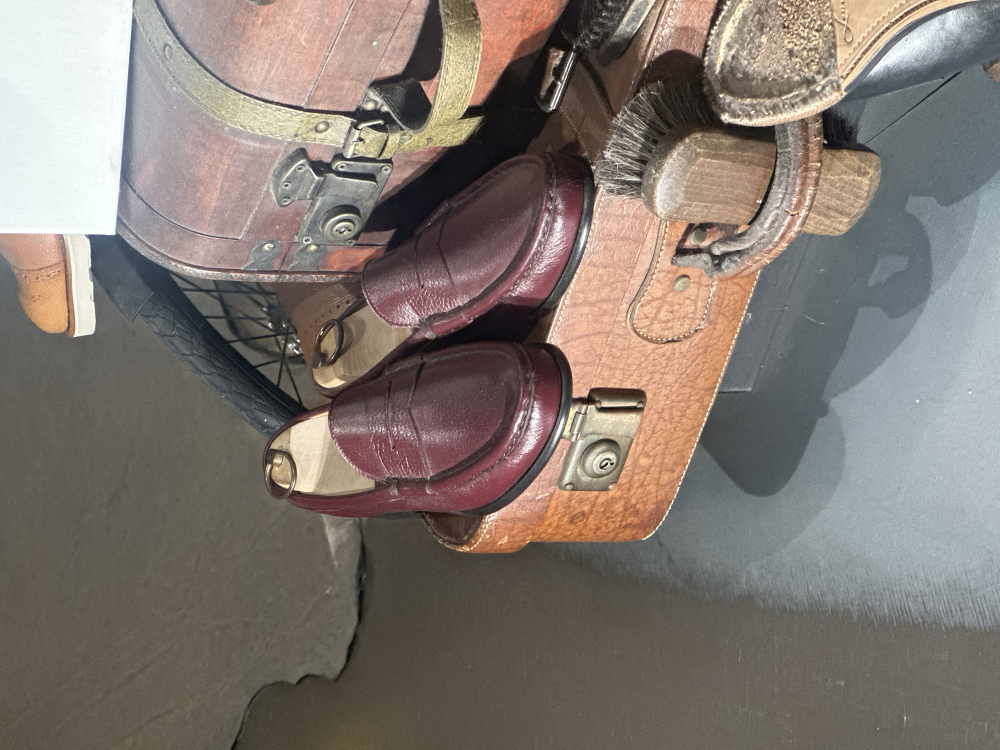 The image presents a close-up, rotated view of several items arranged together, likely as part of a display. The central subjects are two pairs of shoes, two suitcases, and a shoe brush.
One pair of shoes consists of dark red, possibly oxblood, leather loafers with a classic penny loafer saddle strap. They appear to be on shoe trees or forms, maintaining their shape. The other visible shoe is a tan or light brown leather wingtip style, with a thick white sole, peeking in from the upper left.
Beneath and around the shoes are two suitcases or trunks. One is a rich, dark brown, possibly wooden, with a rounded top and secured by olive green fabric straps with brass buckles and metal clasps with keyholes. The other suitcase, on the right, is made of lighter brown, textured leather with visible stitching and a brass-colored clasp.
A wooden shoe brush with dark bristles is tucked between the lighter brown suitcase and the dark red loafers. Part of a black bicycle tire and spokes are visible in the background, near the dark brown trunk. The background also includes a dark grey or black floor and a partial white surface on the far left.
The objects are arranged tightly within the frame, suggesting a curated setup, perhaps in a store or museum exhibit. The general aesthetic is classic and somewhat vintage, focusing on quality leather goods and travel accessories. The image shows no human activity or specific time of day, and all lighting appears artificial. Although the context provided is Taormina, Italy, there are no visual cues within the image to confirm this location. Faint text, possibly a brand name like "Ralph Lauren," is partially visible on the tan shoe.
