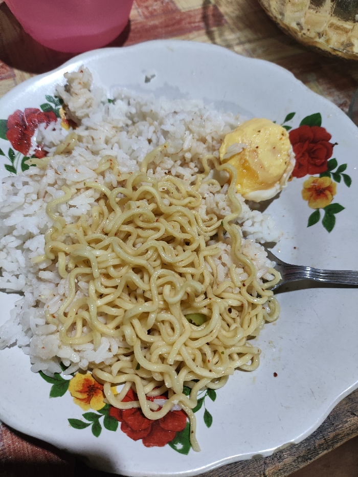 A close-up overhead shot displays a simple meal served on a white floral plate. The plate, which has a scalloped edge, features a pattern of red, yellow, and green flowers and leaves. The main components of the meal are a generous portion of white rice and a serving of yellow, wavy instant noodles, which are placed on top of the rice. Red specks, possibly chili flakes, are visible among the rice and noodles. To the right side of the plate, a halved boiled egg with a soft, yellowish yolk is present. A silver-colored fork rests on the plate, suggesting the meal is ready to be eaten. In the blurred background, a pink cup is visible in the upper left corner, and a textured, possibly golden or metallic object (perhaps a bowl or decorative item) is seen in the upper right. The surface beneath the plate appears to be a worn, rustic material, possibly wood. No people are visible, and there is no visible text. The image provides a glimpse of a casual meal setting, consistent with daily life in Selagai Lingga, Indonesia.