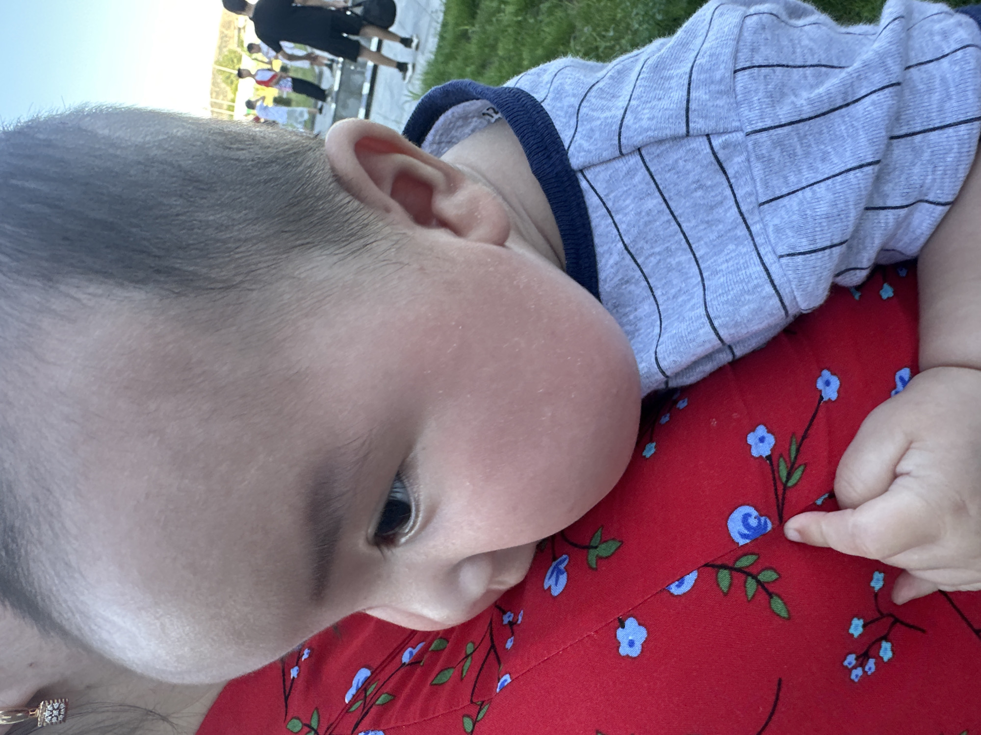 A close-up shot shows a baby being held. The baby's head and face are prominently featured, with the infant's left ear and cheek pressed against the shoulder of the person holding them. The baby is wearing a gray striped long-sleeved shirt with a navy blue trim. The person holding the baby is wearing a red shirt with a blue floral pattern. A finger is visible on the lower right of the frame, resting on the red shirt. In the background, out of focus, several people are standing on a paved area near a grassy lawn. The setting appears to be outdoors, possibly in a park or public space. There is no discernible specific location within the city of Manas, Kyrgyzstan, that can be identified from the visual information provided.