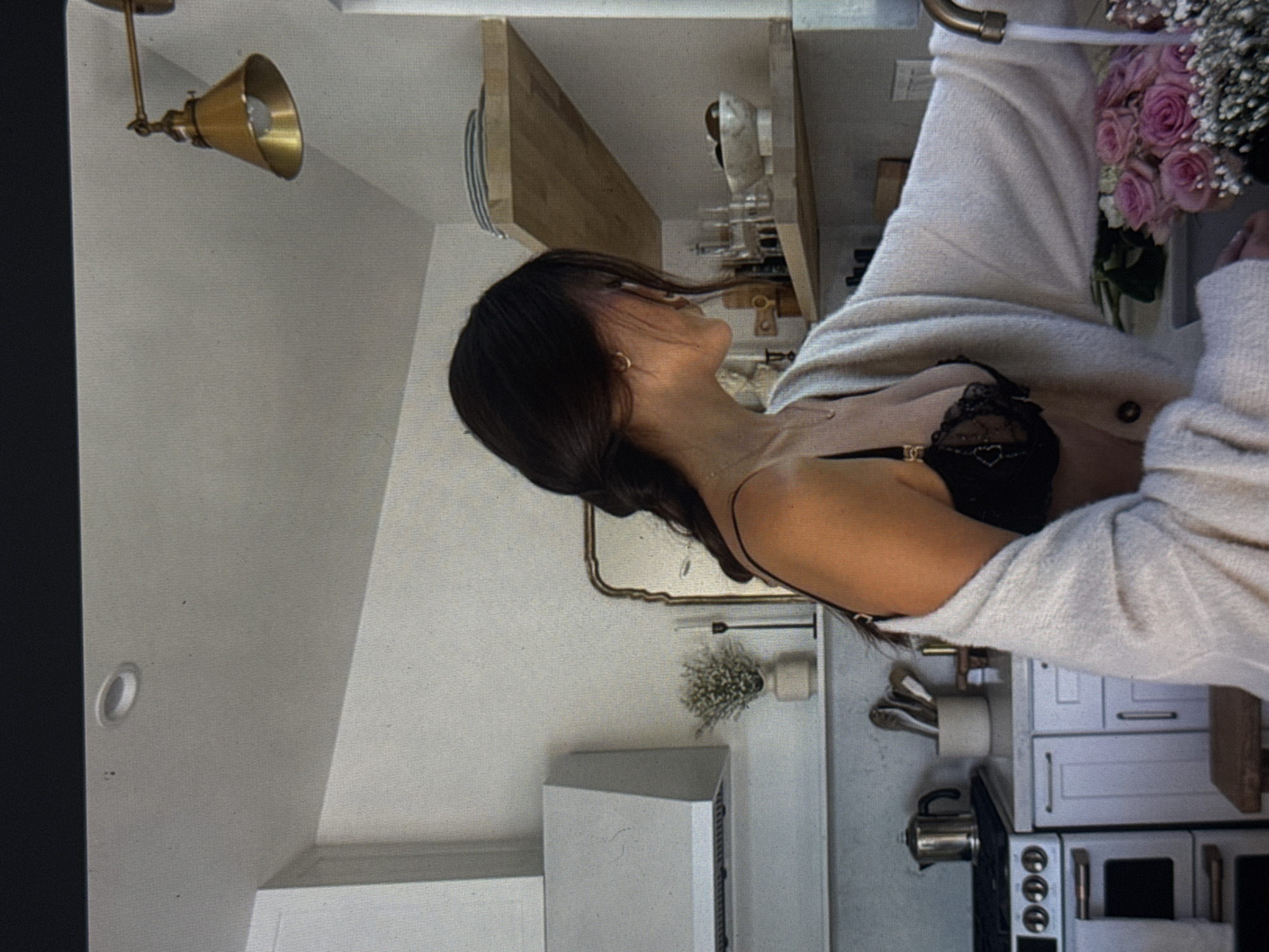 A woman with dark hair in a braid is standing in a kitchen, leaning over a sink. She is wearing a black lace bra and a cream-colored cardigan. A faucet is running water into the sink, where a bouquet of pink roses and baby's breath is placed. A stainless steel range and hood are visible in the background, along with white cabinets and wooden shelves. A brass wall sconce is mounted on the wall above the shelves. The scene appears to be in a residential kitchen. There is no indication of the location being in Kuta Selatan, Indonesia.