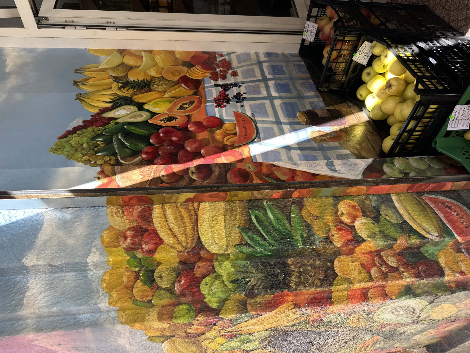 The image captures a fruit stand or market scene, presumably in Sofia, Bulgaria. In the upper part of the frame, a mural depicting a vibrant array of fruits such as bananas, pineapples, grapes, watermelons, and other various fruits is visible. The mural is painted on a wall, possibly of a building or a storefront.
To the right, two black plastic crates are stacked, filled with apples. One crate contains red apples, while the other contains yellow-green apples. There are tags attached to the crates, probably indicating pricing or other relevant information.
The lower part of the image shows the same fruit mural. The stairs is showing the fruit mural at different angles. A beam of sunlight falls across the scene, enhancing the colors of the mural and creating contrasting shadows. The overall environment appears to be an outdoor marketplace or shopfront.