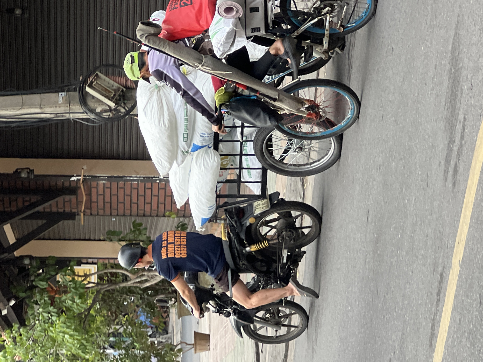 In Nha Trang, Vietnam, two individuals are engaged in transit on the street. The individual on the left operates a black motorcycle, wearing a dark blue t-shirt with orange lettering and a black helmet. The individual on the right pilots a bicycle, dressed in a purple and black long-sleeved shirt, a lime green cap, and black pants, carrying a fishing rod and a red bag on the back. Both are transporting substantial loads of white sacks. The environment includes buildings with roll-up doors and brick facades, foliage, and street infrastructure.