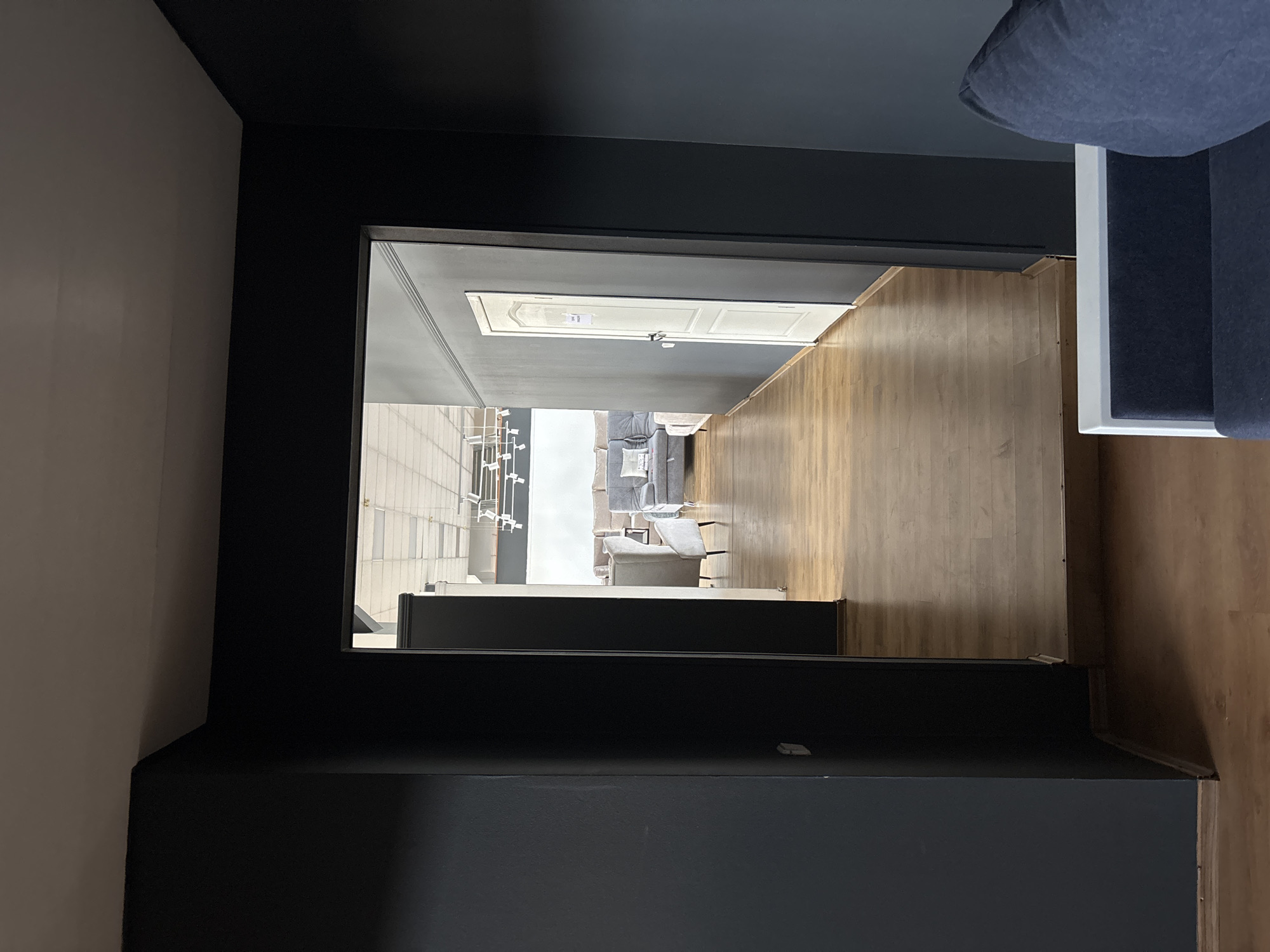 The image shows a view through a doorway into a furniture store. The foreground is dark, with a dark gray wall on the left and a glimpse of a dark blue sofa on the right. The doorway opens into a well-lit showroom with light-colored walls and a wooden floor. The showroom features several sofas and armchairs, and a white door is visible on the right wall. Overhead lighting fixtures are also visible. The scene appears to be an interior view of a commercial establishment, possibly a furniture showroom, located in the city of Zaporizhzhia, Ukraine. There are no people visible in the image.