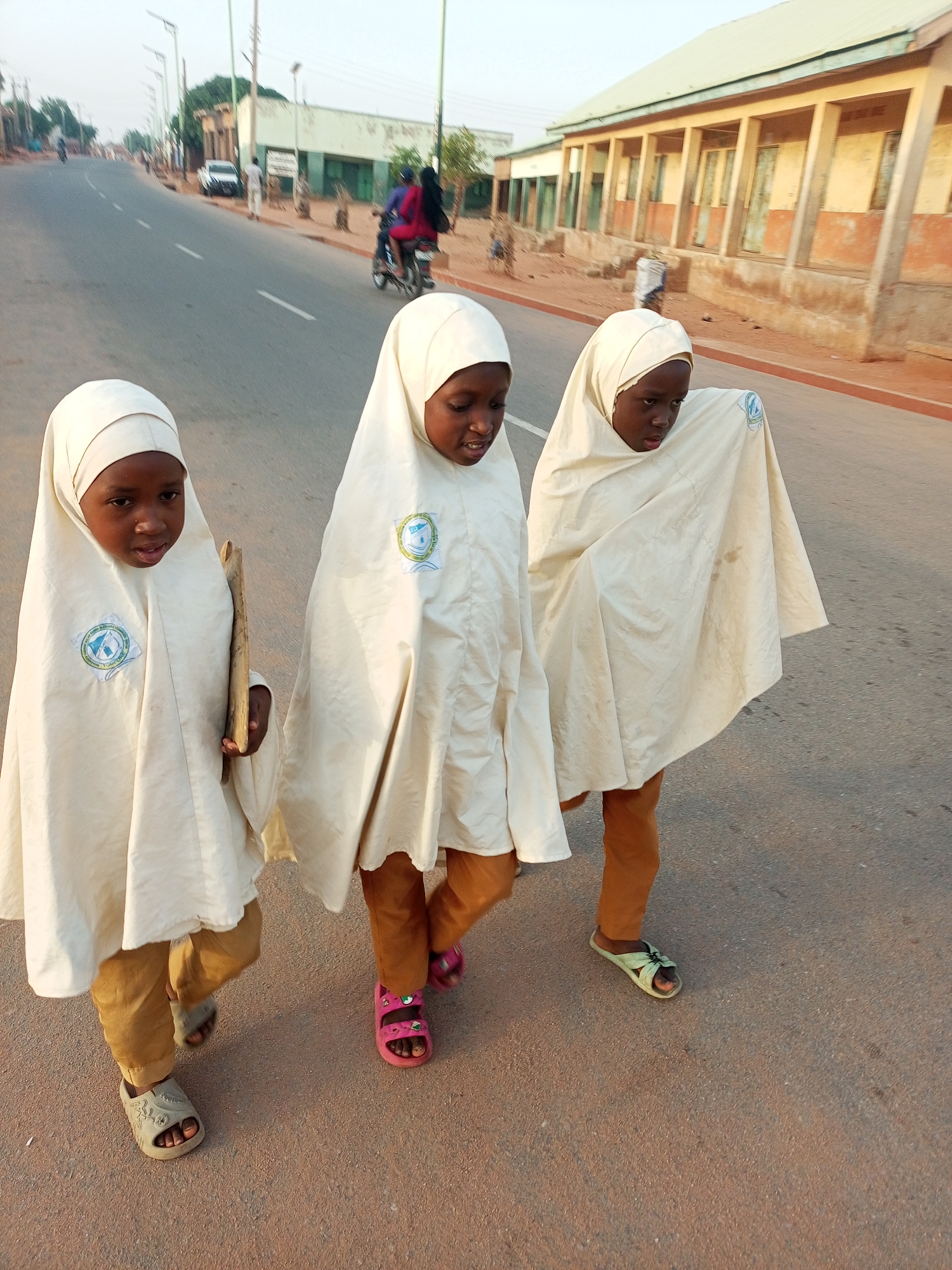 Three young girls, dressed in matching cream-colored headscarves and cloaks over brown pants, are walking side-by-side on a paved road. The girl on the left carries sticks, and each girl wears different colored sandals. A building with a colonnade is visible on the right side of the road. In the background, other vehicles and figures are visible on the road. The location is likely Tempure, Nigeria.