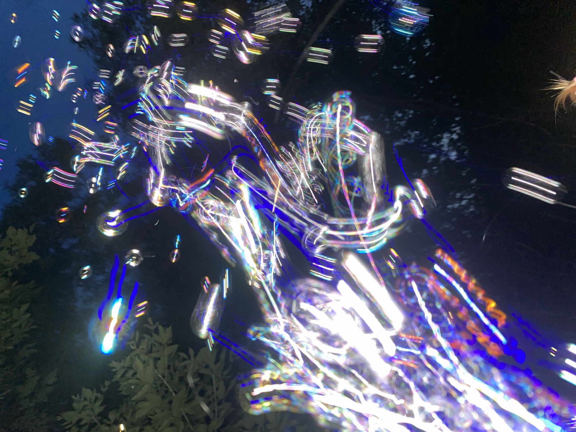 The image depicts a dark, outdoor scene at night, possibly in Novosibirsk, Russia, with numerous illuminated bubbles floating in the air. The bubbles are captured with motion blur, creating streaks of light in various colors, including white, blue, purple, and orange, against a dark background with faint silhouettes of trees and foliage. Some of the bubbles appear to be reflecting light, creating iridescent patterns. There are also a few elongated, cylindrical lights visible, suggesting artificial lighting elements within the environment, possibly part of a performance or attraction. A small portion of a person's blonde hair is visible in the bottom right corner, indicating human presence. The overall impression is one of a vibrant, dynamic, and somewhat abstract display of light and motion.