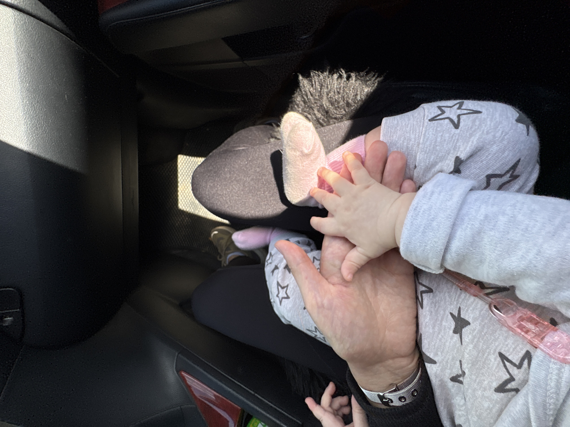 The image shows a close-up view from inside a car. A person is holding a baby in their lap. The baby is wearing gray pajamas with star patterns. The person's hand is large and fair-skinned, wearing a silver bracelet. The baby's tiny hand is reaching over the person's hand. The baby's foot is visible, wearing a pink sock and a light-colored shoe. The car interior is mostly black plastic. It is not possible to determine if this is in Votkinsk, Russia, without further information.