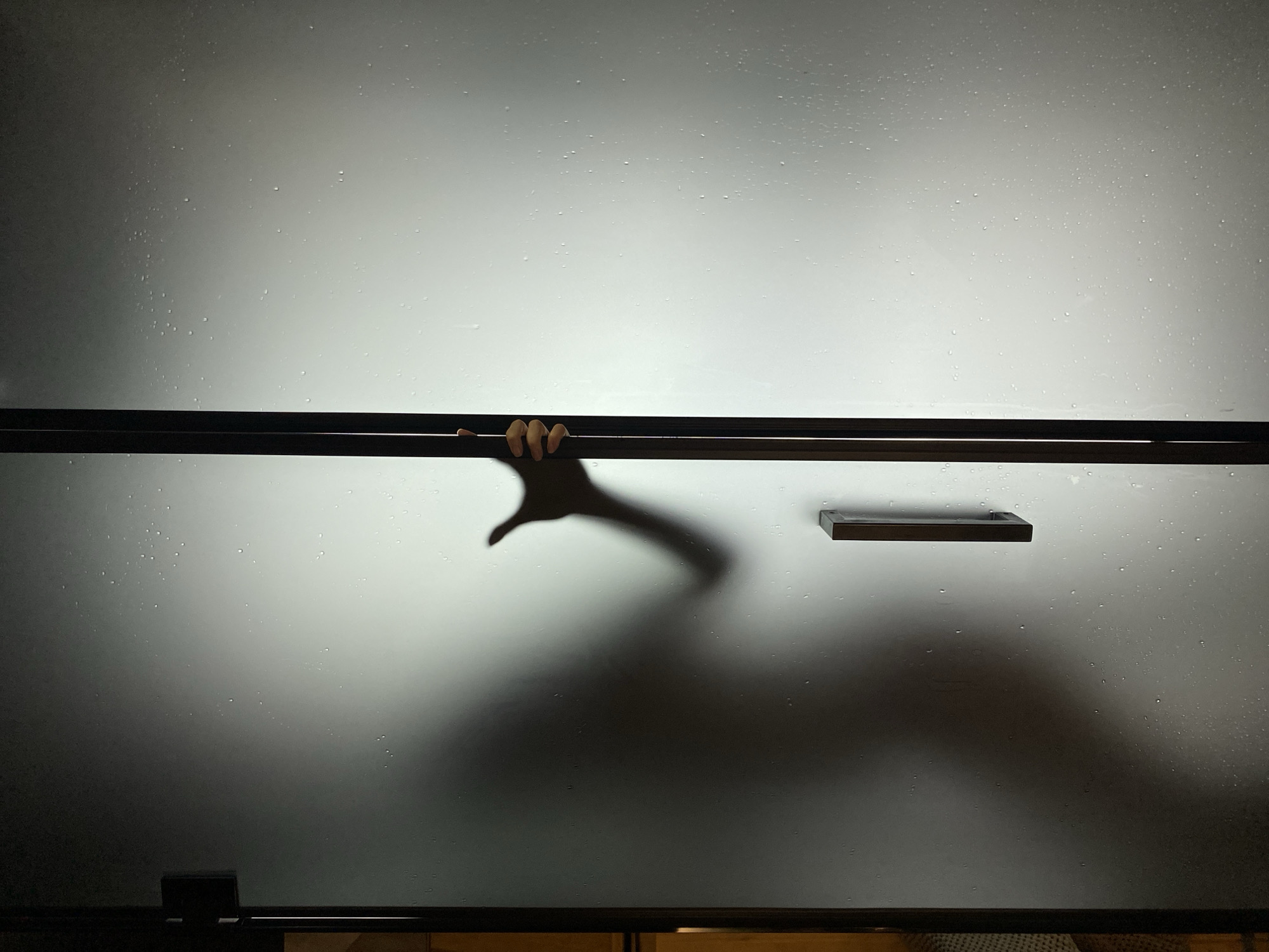 A frosted glass shower door is partially open, revealing a silhouette of a hand reaching for the handle from the other side. The door has water droplets on its surface and a dark vertical handle. A dark shadow is cast on the door's surface, obscuring the individual behind it. The scene appears to be in a bathroom. The image does not contain any elements that can identify its location within the city of Votkinsk, Russia.