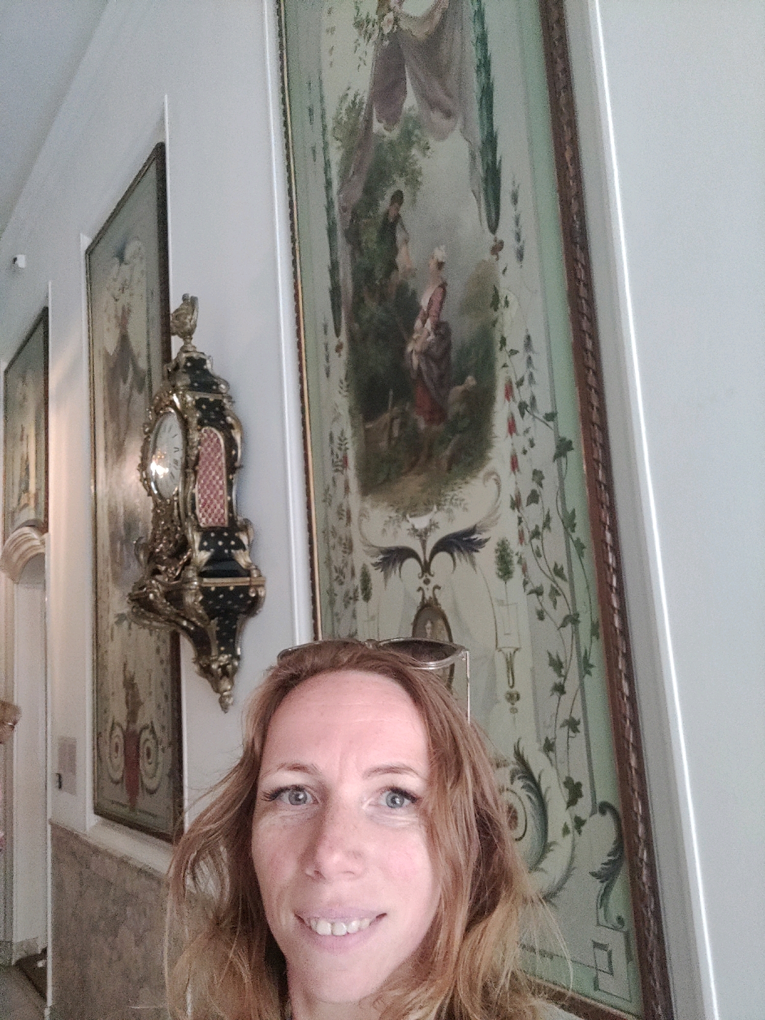 A woman with shoulder-length, wavy blonde hair and blue eyes is captured in a selfie. She is wearing sunglasses pushed up on her head and smiling. The background features ornate, traditional artwork hanging on a light-colored wall. To her left, a vintage clock with intricate gold detailing and a dark enamel casing is mounted on the wall. The artwork includes pastoral scenes and decorative floral motifs. The scene suggests an indoor setting, possibly a museum or a historic home. The overall impression is a personal snapshot taken during a visit to a culturally rich location. This image is located in the Rijksmuseum in Amsterdam, Netherlands.