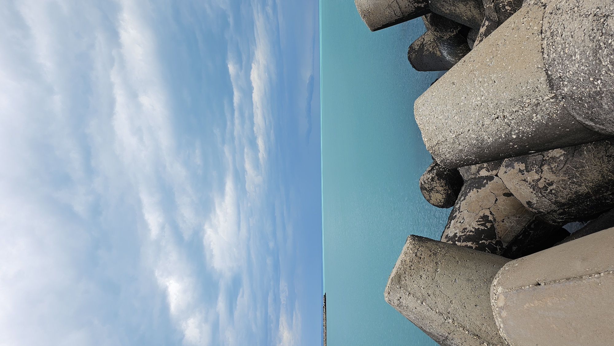 The image shows a view of the sky, sea, and breakwater in Varna, Bulgaria. The left side of the image is dominated by a bright blue sky with scattered white clouds. The right side of the image features a section of the sea, which appears as a bright turquoise color. Along the edge of the sea are several large, gray concrete tetrapods forming a breakwater. The tetrapods are arranged haphazardly and show signs of weathering and age.