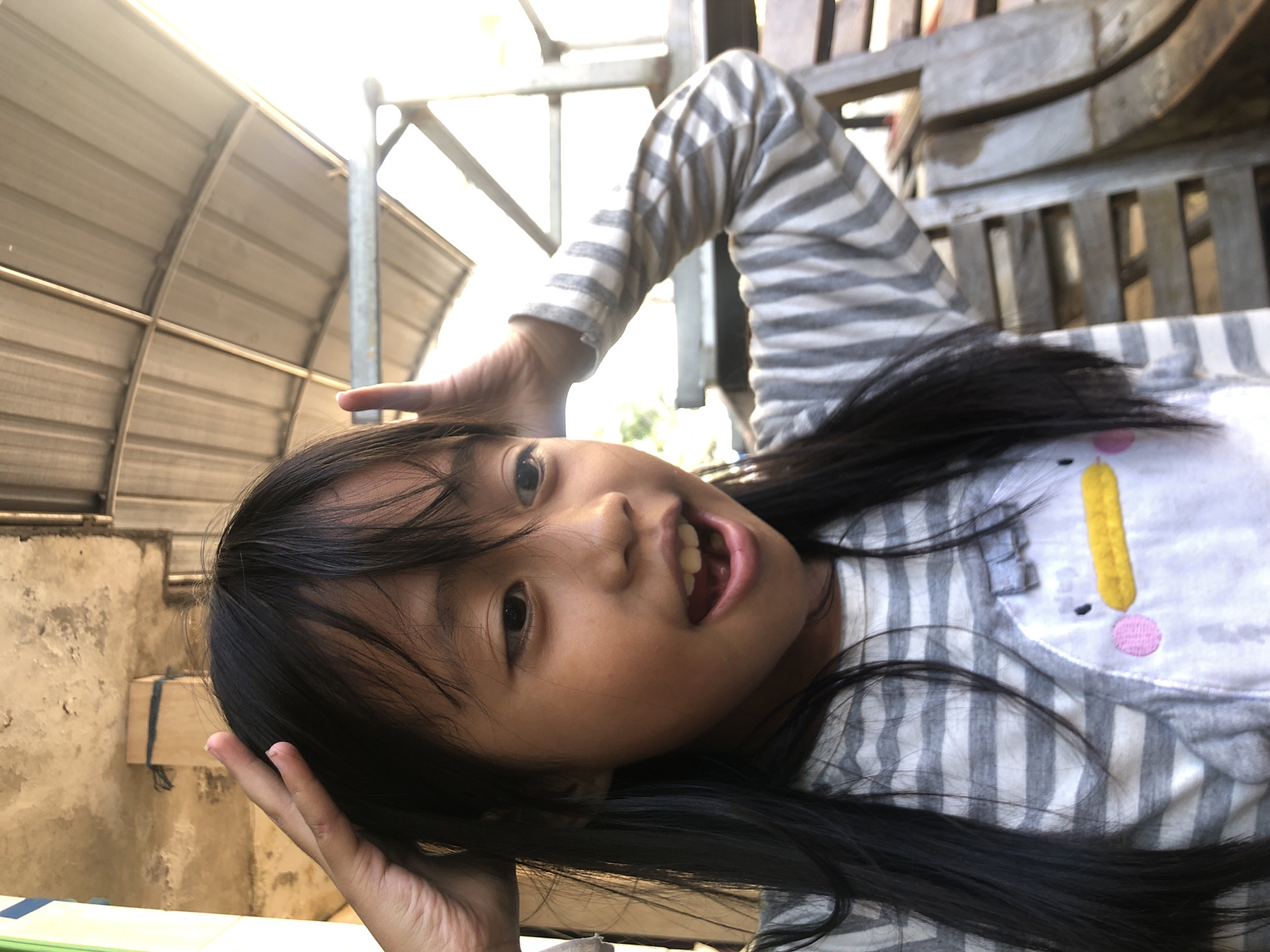 A young girl with long, dark hair is shown in a close-up, medium shot. She is wearing a gray and white striped long-sleeved shirt with a penguin design on the front. The girl has her hands on her head and is looking to her right with her mouth open. The background appears to be an outdoor, covered area with a corrugated metal roof, suggesting a casual setting, possibly a porch or open-air structure. Wooden furniture, likely benches, can be seen to the right. The lighting is bright, indicating daytime. The image is taken from a slightly low angle, looking up at the girl. The overall impression is that of a playful or surprised moment captured.