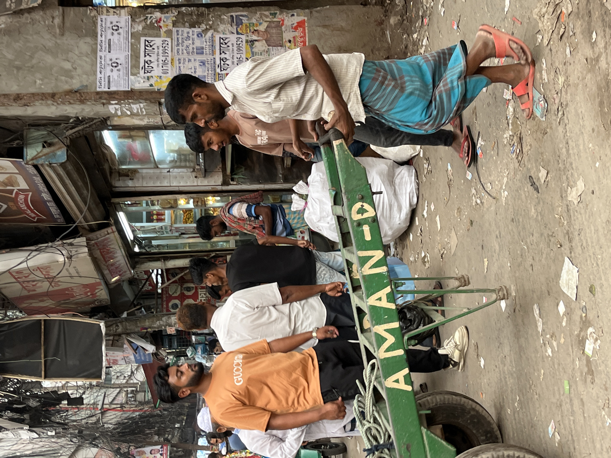 The scene depicts a crowded street in Dhaka, Bangladesh. Several people are seen navigating the narrow space, pushing a green handcart with "AMAN-D" written on it. The cart appears laden with white sacks. Two men, one wearing a checkered lungi and an older shirt and the other in a beige shirt, are pushing the cart, while another man in an orange "GUCCI" T-shirt and black pants follows behind. Additional people surround the cart, some dressed in t-shirts and others in more traditional attire. The background features densely packed buildings, posters, and overhead power lines, indicative of the city's infrastructure. The street surface is littered with debris and discarded paper.
