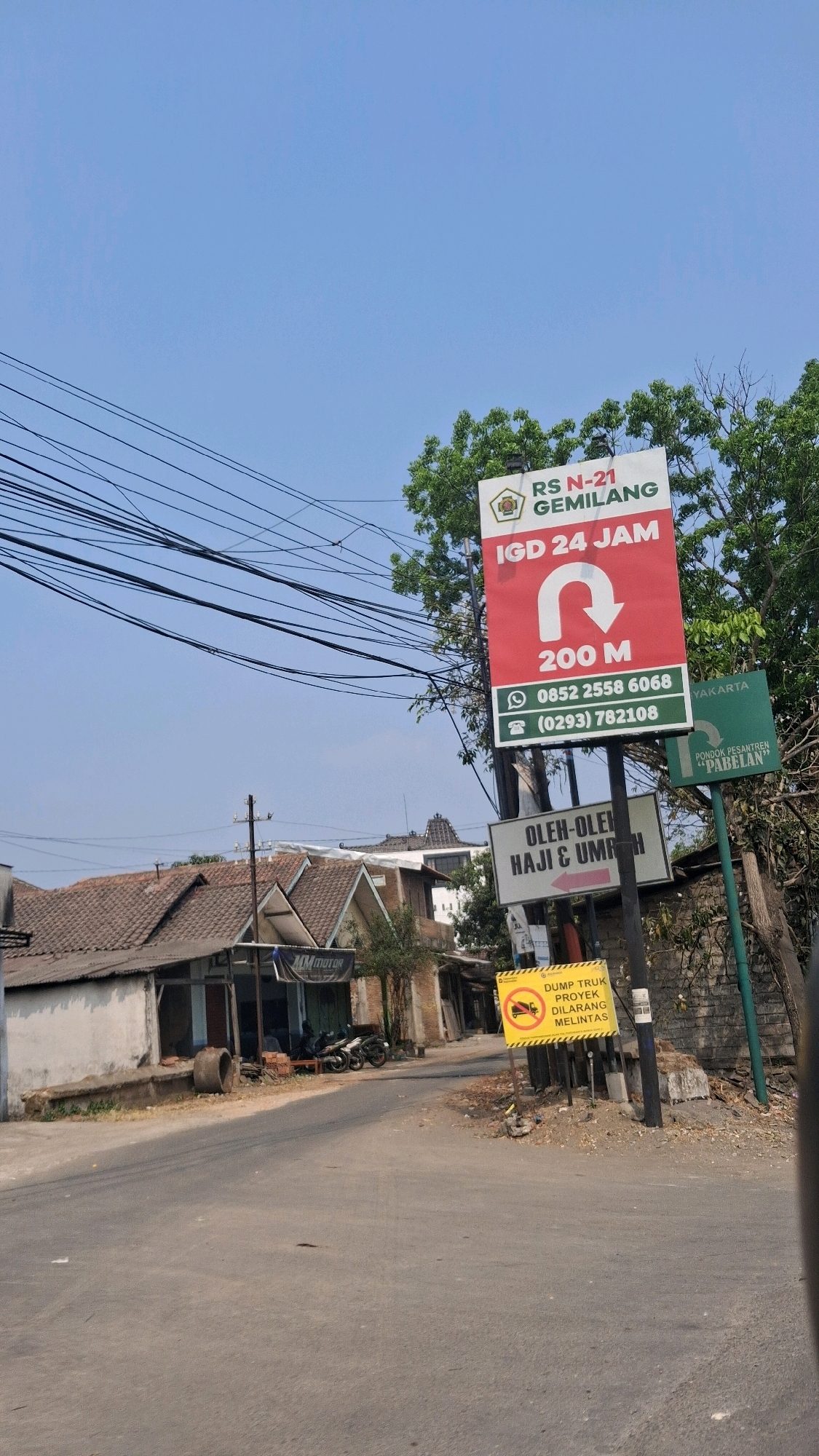 The image captures a street scene in Pabelan, Indonesia, under a clear blue sky. A prominent red and white sign indicates "RS N-21 GEMILANG IGD 24 JAM" with a U-turn arrow and "200 M". Below this, a smaller white sign reads "OLEH-OLEH HAJI & UMROH" with a left-pointing arrow. A yellow sign with a red circle and slash over a truck symbol states "DUMP TRUK PROYEK DILARANG MELINTAS".
To the left of the road, several low-pitched tiled-roof buildings are visible, one with a sign for "MM Motor". A few motorcycles are parked in front of one building. The road itself is paved and appears to be a typical Indonesian street with overhead power lines stretching across the frame. The environment is somewhat rural or suburban, with trees and vegetation present, suggesting a warm climate. No people are clearly discernible in the image.