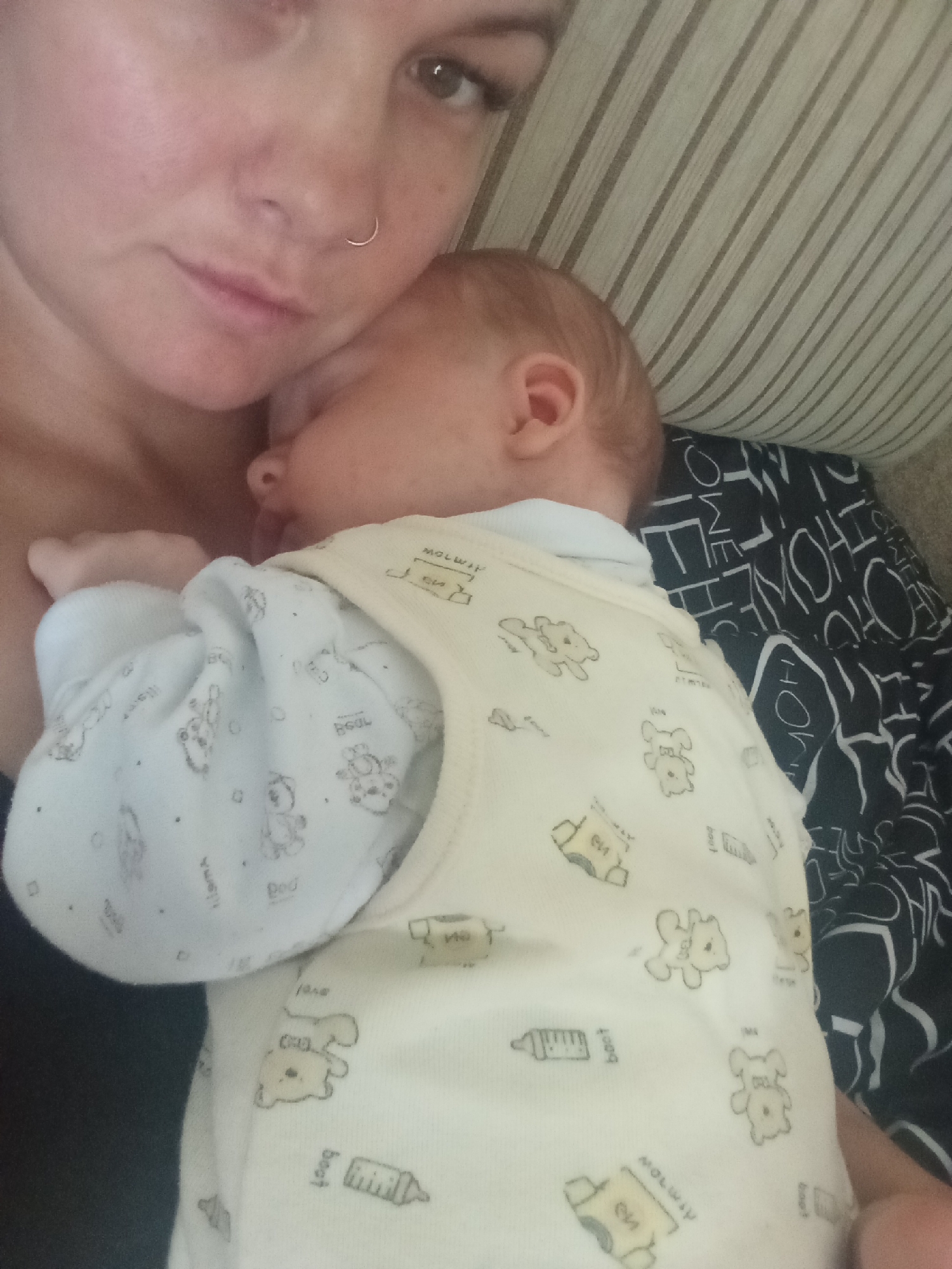 A close-up selfie shows a woman holding a sleeping baby. The woman is on the left side of the frame, her face partially visible as she looks down. She has a nose ring. The baby is nestled in her arms, facing away from the camera. The baby is wearing a light-colored onesie with a pattern of teddy bears and baby bottles. The background consists of a striped cushion and a black and white patterned fabric. There is no information in the image to suggest a location in Chaikovsky, Russia.