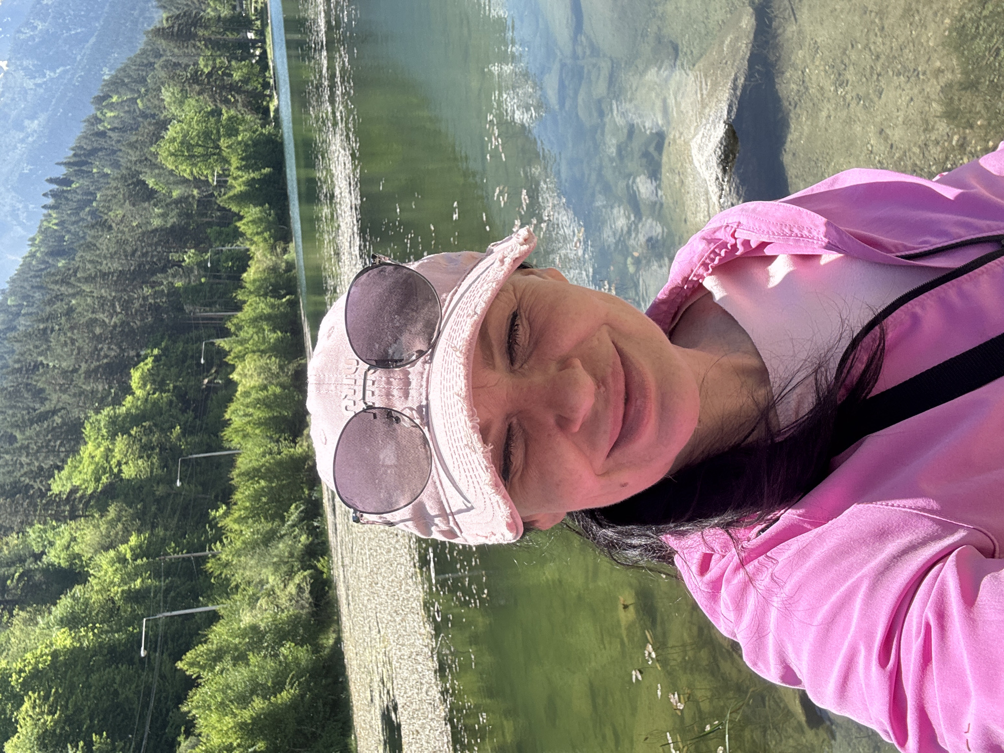 A woman wearing a pink baseball cap and a bright pink jacket takes a selfie by a body of water. Her eyes are closed, and she is smiling. Sunglasses are perched on the brim of her cap. The water is green and has lily pads floating on its surface. In the background, a lush green forest covers a hillside, with tall trees reaching towards a clear blue sky. The scene appears to be in a natural outdoor setting, possibly a lake or a calm river, at the city of Teberda, Russia.