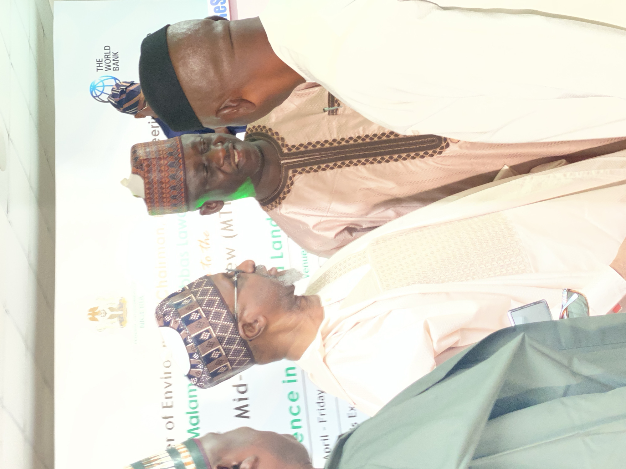 The image captures a scene from an event held in Kwarau, Nigeria. Three men are prominently featured in the frame. Two of the men are facing towards each other. The man on the left is dressed in a traditional attire with a patterned cap, while the man on the right is wearing a black cap and a patterned garment. Behind them, part of a banner is visible, mentioning “er of Enviro”, "Malaman", "Mid-", and "ence in". The banner also displays the logo of the World Bank and the coat of arms of Nigeria. Other text fragments, such as "To the," "Land," and "Venue" can be seen on the banner, but are obscured. The date "April - Friday" is also written, suggesting the timing of the event. The lighting of the image is bright, possibly due to indoor artificial lighting. The image seems to be taken at an angle, likely by someone standing to the side.