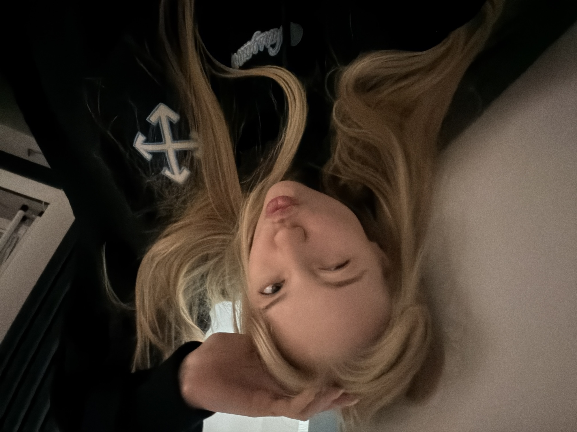 A blonde-haired person lies on a white surface, possibly a bed, with their head tilted. They are winking and pouting. They are wearing a black garment with a white cross emblem. The location is Unknown, Unknown.