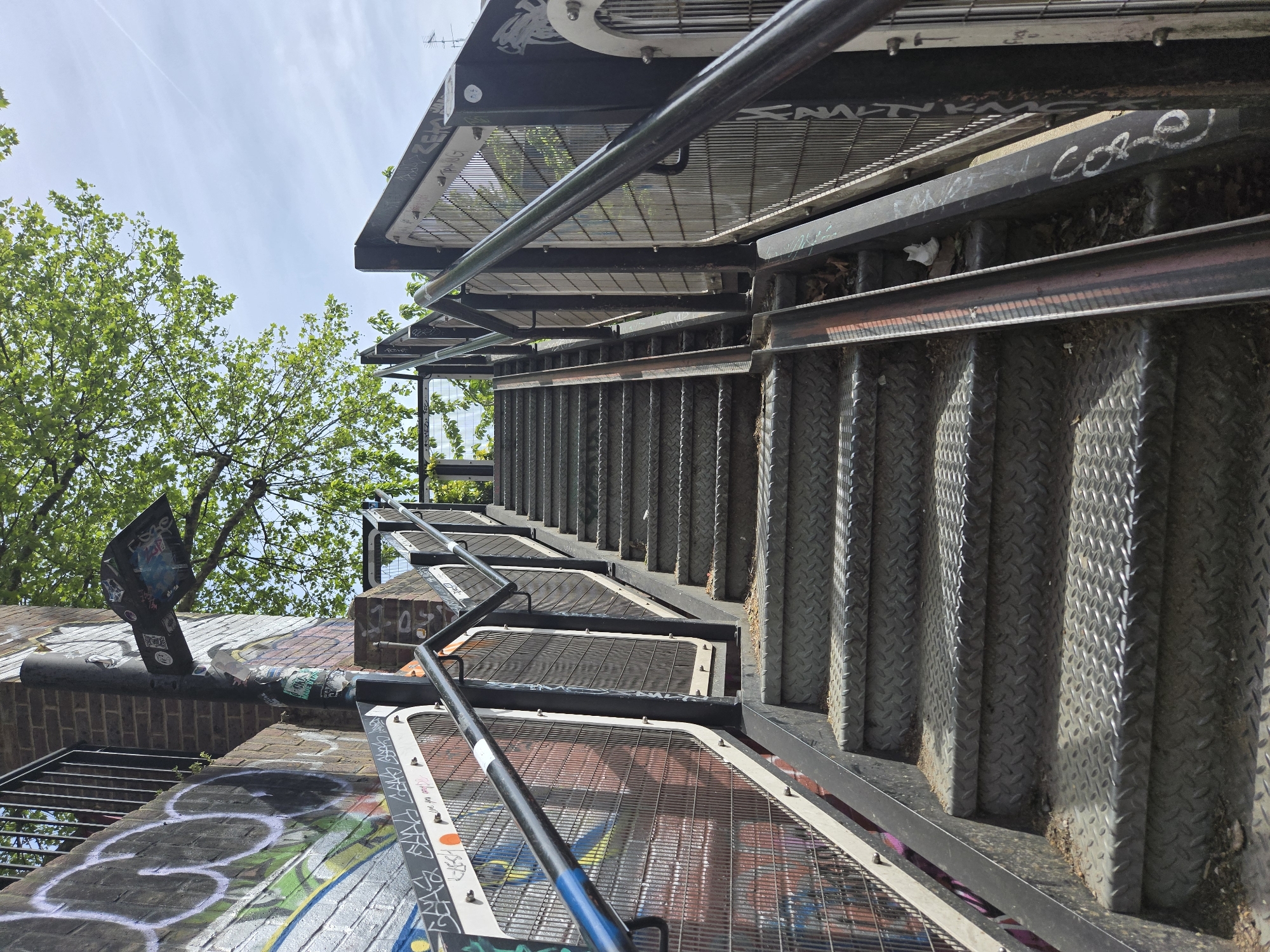 The image depicts an outdoor staircase structure in an urban setting, possibly London, United Kingdom. The staircase is made of metal and has a unique design, with diamond-patterned steps and metal railings. The structure is covered in graffiti, indicating its presence in a public or semi-public space. A brick wall with additional graffiti is visible at the base of the structure. The sky is partly visible in the background, along with the green foliage of trees.