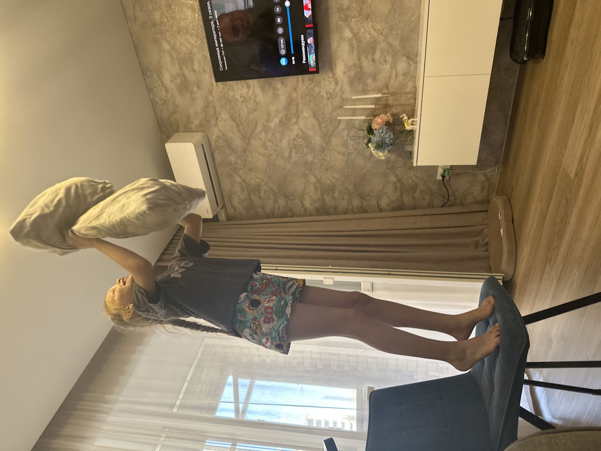 In a bright, modern living room in Abakan, Russia, a young girl with blonde hair braided down her back is captured mid-action. She stands on a dark blue chair, her legs extended as if balancing or performing a playful stunt. In her hands, she holds two light-colored pillows, one above the other, suggesting a possible pillow fight or a moment of imaginative play. Her expression is one of focus and lightheartedness.
The room features a large television mounted on a textured, light gray wall, displaying text in Russian, including "Сокровища императора, 3 сезон" and "Рекомендуем." Below the TV, a white cabinet stands against the wall. An air conditioning unit is mounted high on the wall to the left. The floor is wooden, and sheer curtains are visible in the background, suggesting it might be daytime. The overall atmosphere of the image is casual and domestic, hinting at a relaxed afternoon at home.