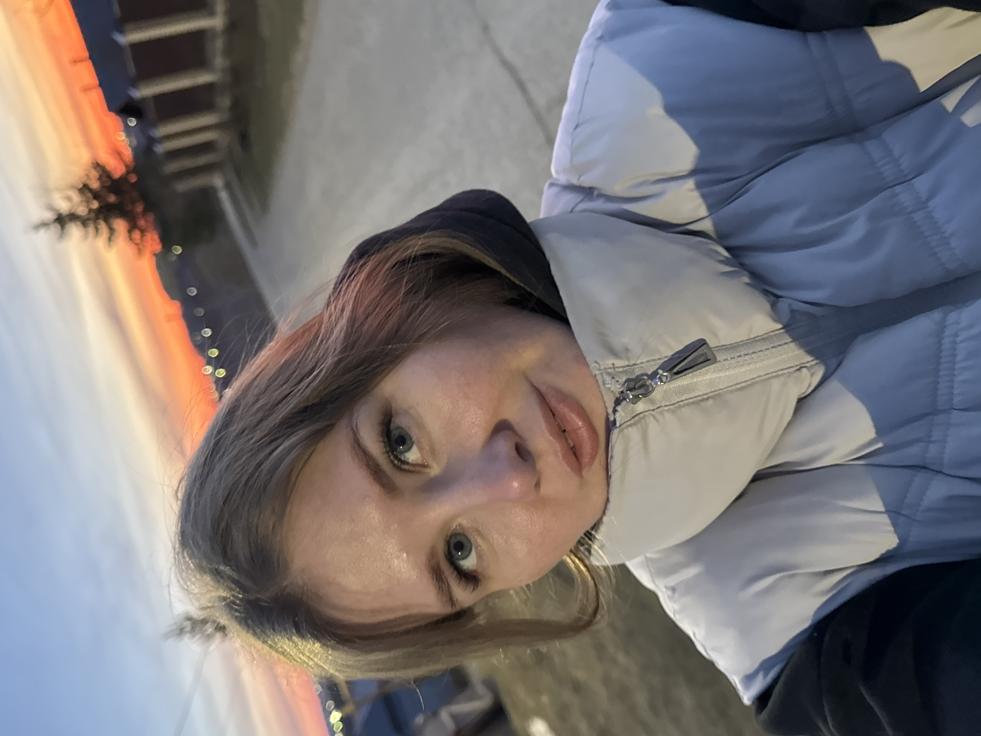 The media file is a close-up, slightly tilted selfie of a young woman taken outdoors during twilight in Горный, Russia. The woman is positioned centrally, looking directly at the camera with a calm expression. She has striking blue-green eyes and light brown hair with subtle highlights. She is wearing a light-colored (possibly pale blue or light grey) puffer jacket with a visible metallic zipper detail on the collar, layered over a dark hoodie or top.
The background is highly blurred, creating a shallow depth of field. Warm hues of orange, pink, and yellow dominate the sky on the left side of the frame, characteristic of a sunset or sunrise. Blurred bright spots, likely streetlights or decorative lights, are visible throughout the background. To the left, a dark, indistinct shape that resembles a fir tree or evergreen stands against the colored sky. A muted, paved surface can be vaguely discerned below.
The image conveys a tranquil atmosphere, fitting the serene twilight hour. No visible text is present in the image.