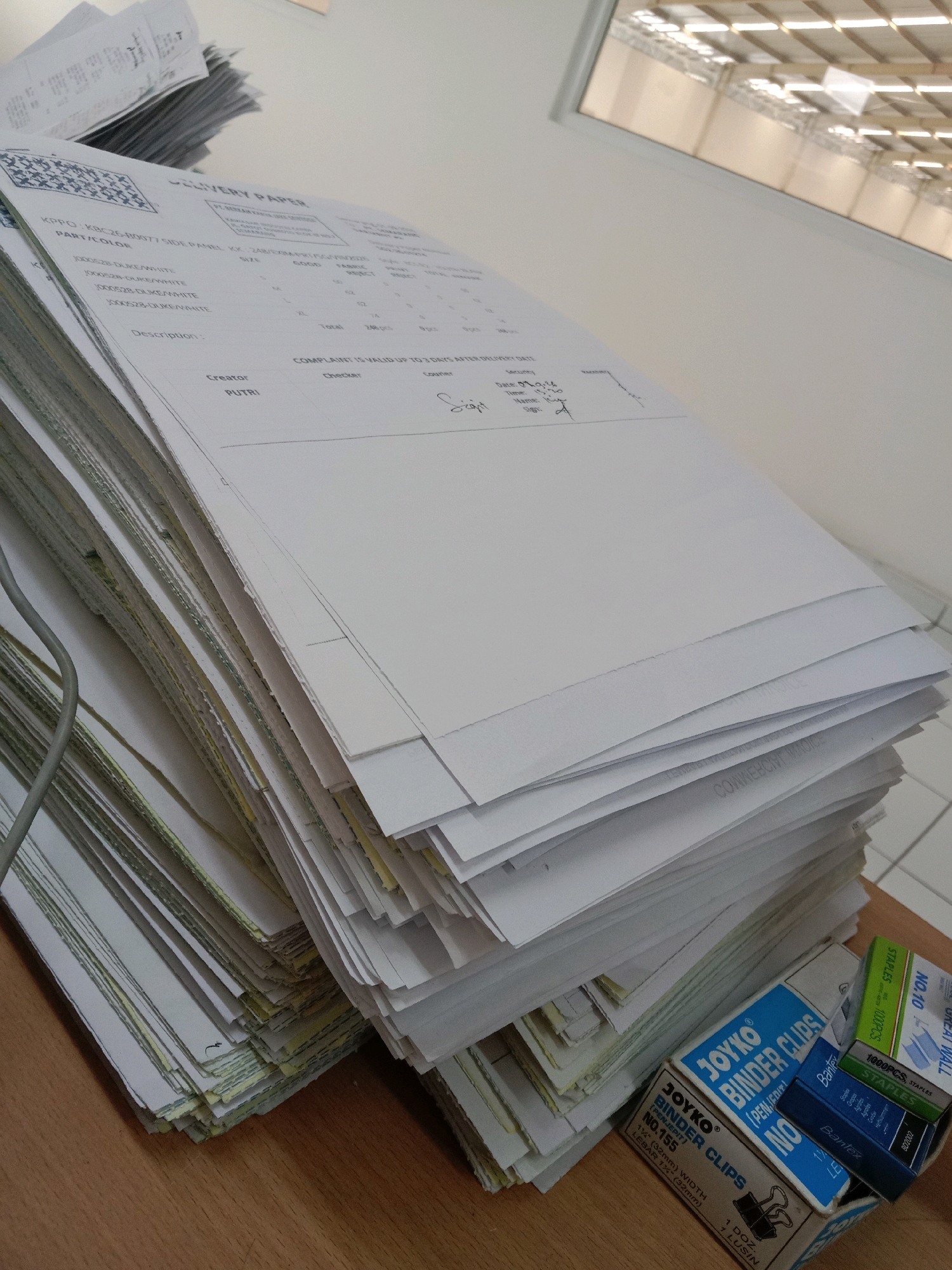 The image shows a stack of papers on a wooden desk. The papers appear to be delivery slips or invoices, with some handwritten information on them. In the foreground, there are several boxes of office supplies, including staplers and binder clips. The background shows a window with a view of a building with a large skylight, possibly a warehouse or factory. The overall impression is that of an office or storage environment. The image does not contain any elements that would allow for the determination of the specific location within Semarang, Indonesia.