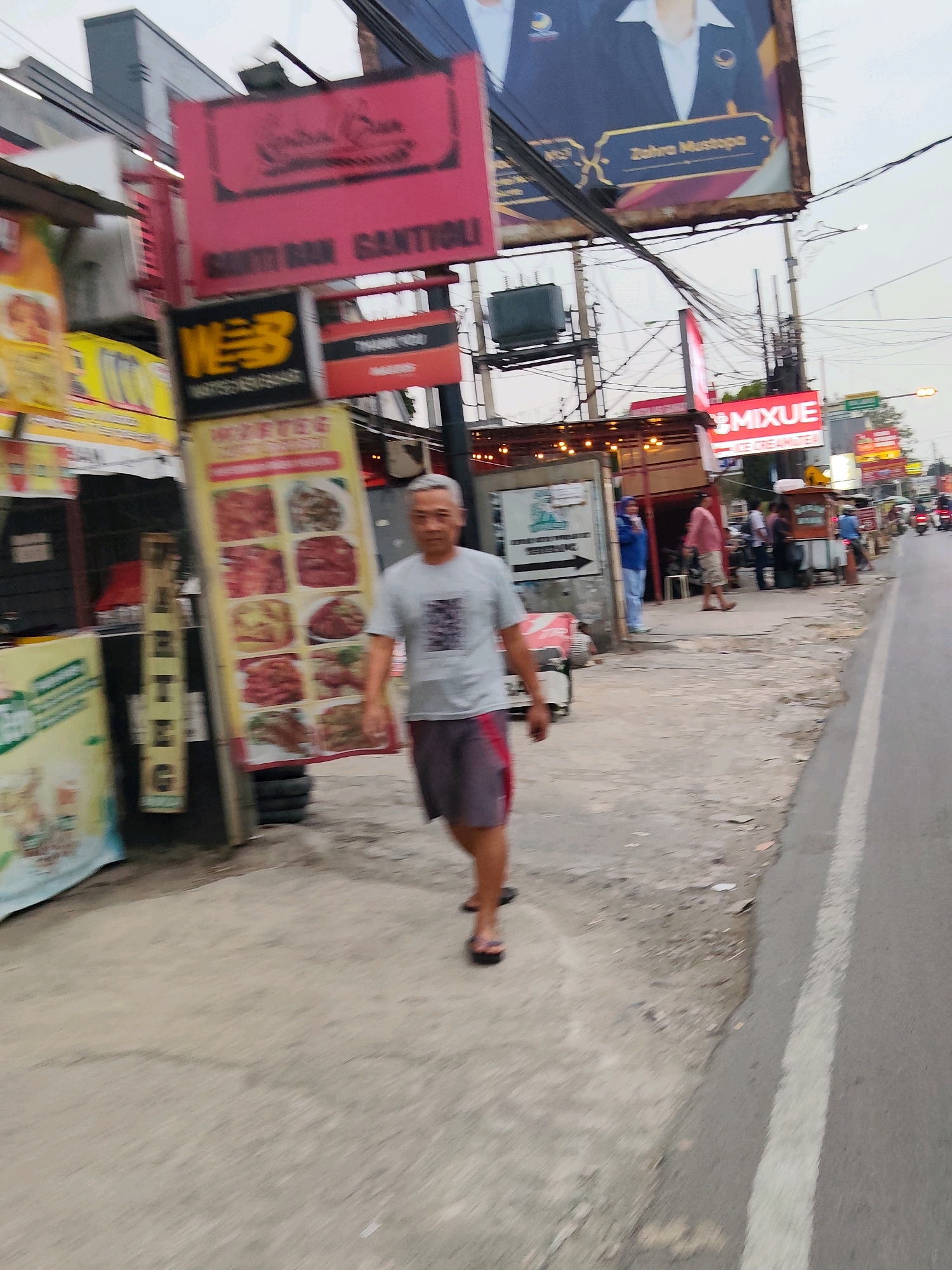 A man in a grey t-shirt and grey shorts with red trim walks on a concrete sidewalk beside a busy road in Purwakarta, Indonesia. He is walking away from the camera. Several signs are visible along the sidewalk, including a large red sign with text in black, a sign with the word "WEB" in yellow and black, and a vertical yellow sign displaying images of food. In the background, there are buildings, other people, and vehicles. The sky is overcast.