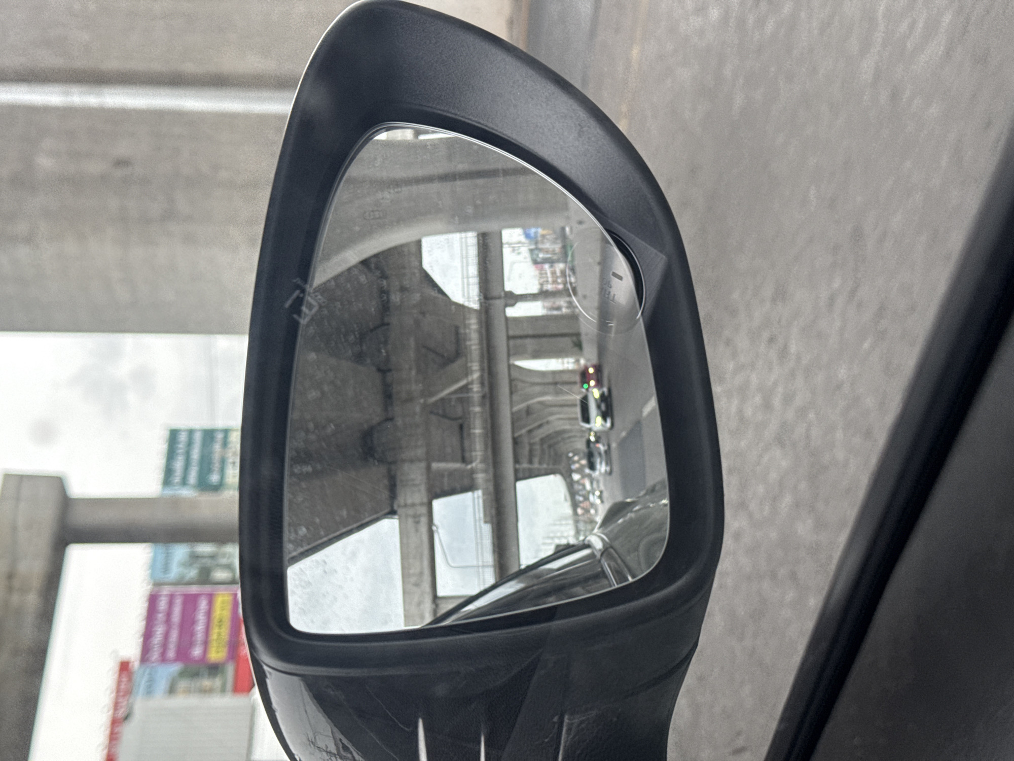 An over-the-shoulder, driver's side perspective shows a reflection in the side-view mirror of a car. The car is driving on a road with multiple lanes, likely an elevated highway or overpass, indicated by the concrete support structures and overpasses visible in the reflection. Traffic is visible in the mirror, with several cars driving in the same direction. In the background of the reflection, billboards can be seen on the side of the road. The sky appears overcast. The scene is consistent with traffic in an urban environment, possibly in Bang Nam Chuet Subdistrict, Thailand, given the infrastructure and billboard style, though no definitive landmarks are present.