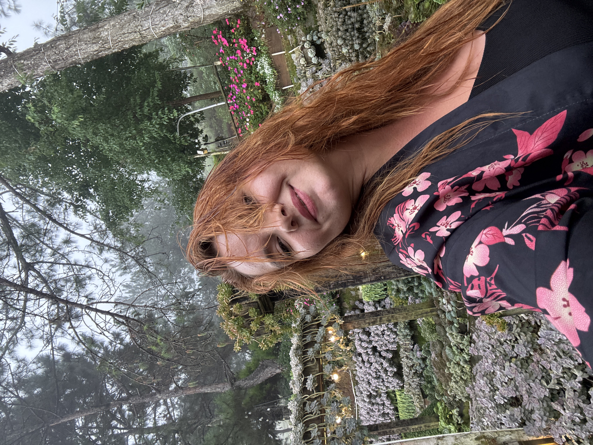 A woman with long, red hair is taking a selfie outdoors in a garden. She is wearing a black shirt with a floral pattern of pink flowers. The background shows lush greenery, trees, and a variety of plants, some of which are in hanging planters. There are also string lights visible in the garden. The setting appears to be a botanical garden or a landscaped outdoor space in Lộc Quý, Vietnam, characterized by its misty atmosphere and abundant flora.