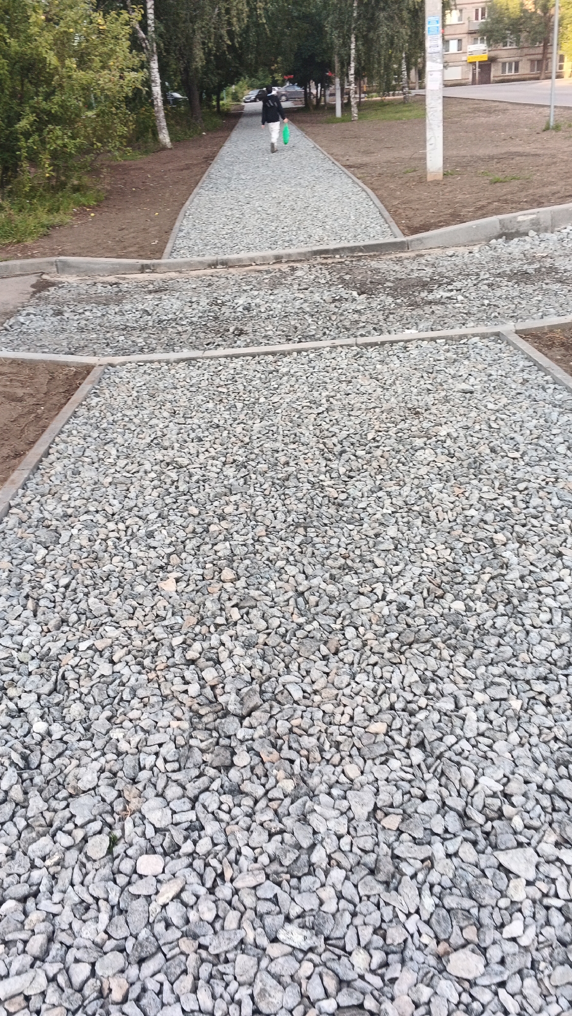 This is a low-angle, full-frame, vertical, eye-level, daytime, outdoor, street-level, wide shot image of a newly constructed or under-construction gravel pathway in a park or urban green space, likely in Izhevsk, Russia, as indicated by the architectural style of the building visible in the background and the foliage present. The pathway is composed of light gray crushed stone, which forms the foreground and extends into the midground. The pathway appears to be bordered by concrete curbs or edging. In the midground, a person is walking away from the camera along the gravel path, carrying a green bag. This individual is wearing a dark jacket and white pants. Beyond the path, trees with green foliage line the area, and a multi-story building with visible windows and some posters on a utility pole are present in the background, suggesting an urban environment. The ground surrounding the path is a mix of dirt and loose gravel, indicating ongoing construction or maintenance.