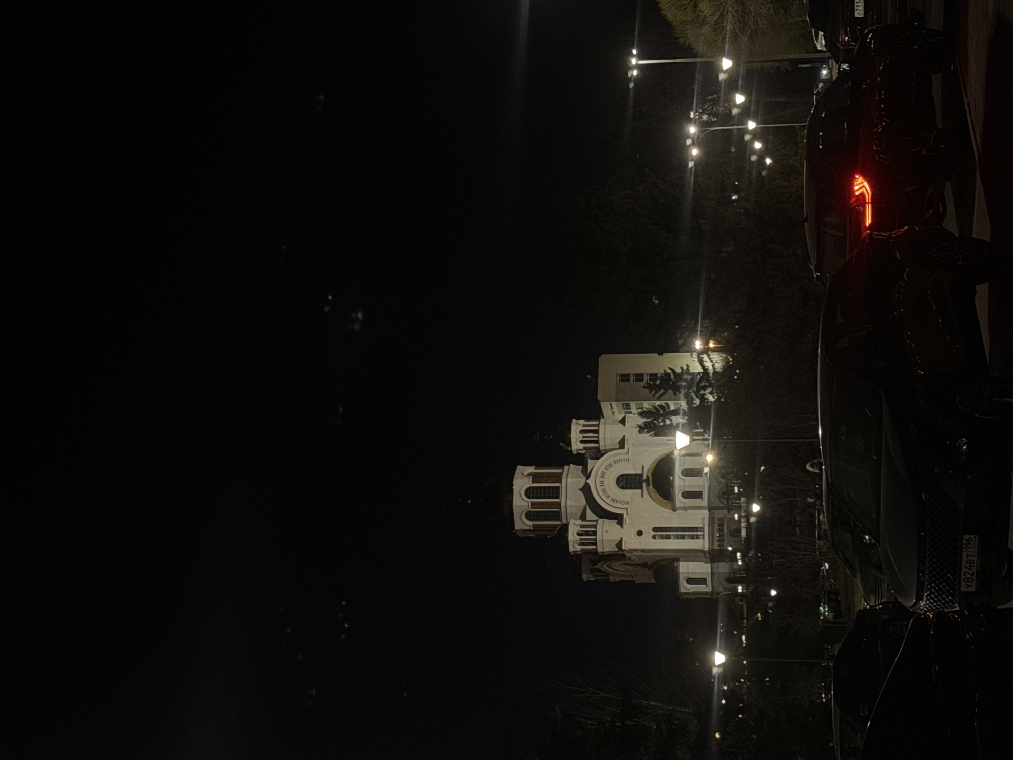 The image depicts a night scene in Yekaterinburg, Russia. A large, white Orthodox church stands illuminated, likely a significant landmark. Streetlights line the area, providing additional illumination. Parked cars are visible along the edge of the frame, indicating a street or parking area near the church. The sky is dark, with minimal ambient light other than the artificial sources. The overall environment suggests a quiet, urban night scene centered around the illuminated church.