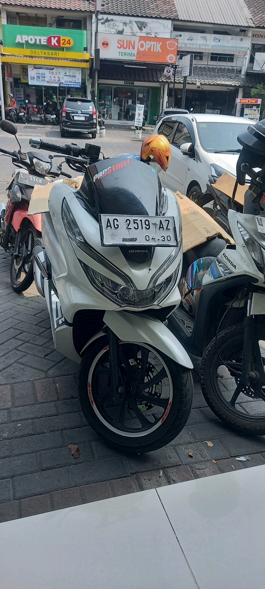 The image shows a street scene in Kureksari, Indonesia, with several motorcycles parked on a brick-patterned sidewalk. In the foreground, a white Honda PCX motorcycle is prominently displayed, featuring a license plate that reads "AG 2519 AZ" and a small sticker with "04.30". A portion of another white Honda motorcycle is visible to the right of the PCX. To the left, a red motorcycle with license plate "L 5968" is partially visible. In the background, various storefronts line the street, including "APOTEK K 24 DELTASARI" and "SUN OPTIK", suggesting a commercial area. A dark-colored car is parked further down the street. An orange motorcycle helmet rests on the windshield of the white PCX. The overall environment appears to be a sunny day, with shadows cast on the ground.