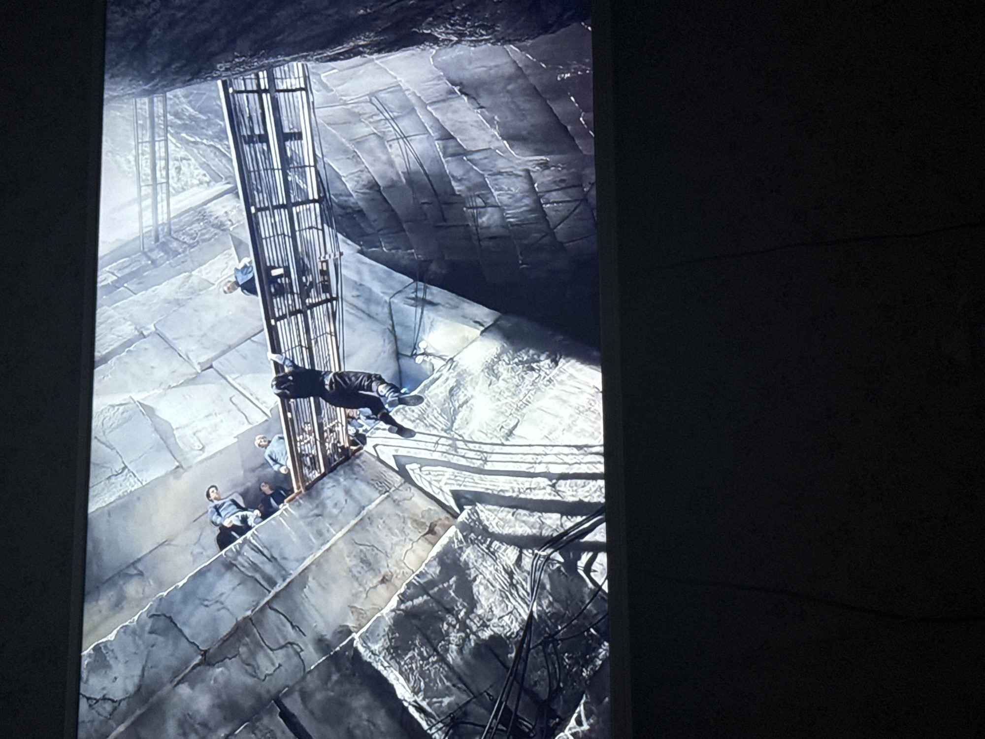 The image depicts a scene from a film or television show, showing a group of people within what appears to be a large, stone structure. The structure is likely a multi-level environment with stairs and a metal cage-like elevator or scaffolding. Several individuals are standing on a lower level, while another person seems to be falling or jumping from the scaffolding above. The environment has a rough, industrial look, with large stone blocks forming the walls and floor. The lighting is dramatic, with strong shadows and highlights adding to the overall depth and mood of the scene. It is not possible to determine the location within the city of Mytishchi, Russia, from the image alone, as the setting is fictional and the structure's characteristics do not resemble any specific identifiable location.