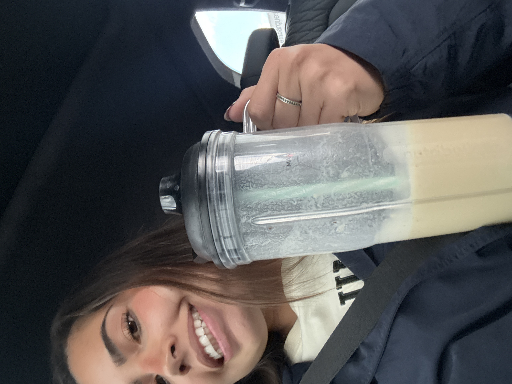 A person with long brown hair, wearing a dark blue jacket and a white shirt with black lettering, is holding a clear blender cup filled with a light tan-colored liquid. The person is smiling and has fair skin. The background appears to be the interior of a car, given the dark interior and a glimpse of the sky through the windshield. The person is wearing a ring on their hand that is holding the blender cup. The location is unknown beyond the information provided in the prompt. There is no indication of Ixtapaluca, Mexico.