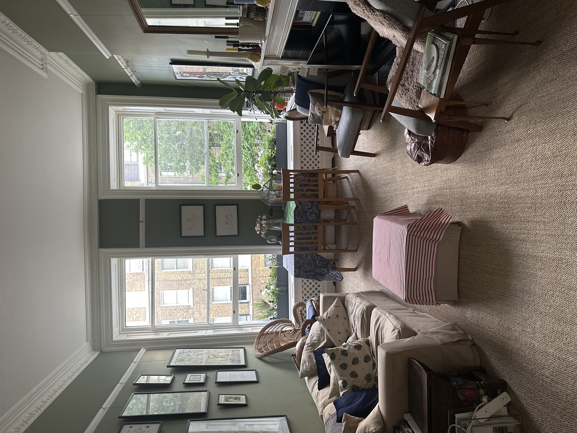 The image shows a room in a building in London, United Kingdom, based on the architectural style of the buildings visible through the windows. The room features two large windows looking out onto an urban street scene. Interior details include a couch with cushions, a coffee table, a dining table with chairs, a fireplace, and an assortment of pictures and artwork on the walls. The room is decorated with a mix of furniture and decor, suggesting a residential setting. There are plants visible in the window boxes. No people are visible in the photograph.