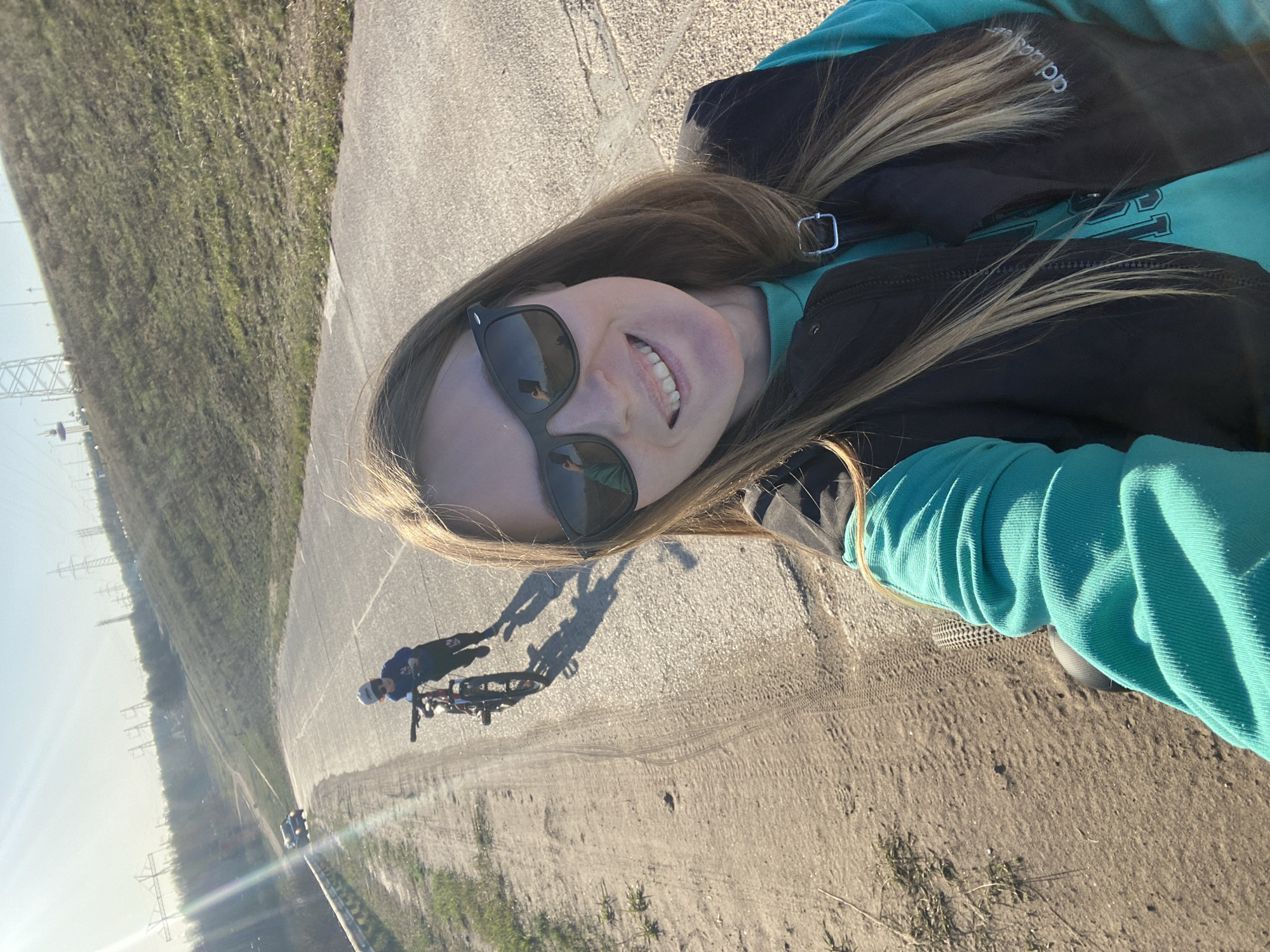 A selfie photo was taken on a paved path in the city of Chaikovsky, Russia, with a blonde woman in the foreground wearing sunglasses, an aqua sweatshirt, and a black vest. The photo is oriented on its side. In the background, a cyclist is visible on the path. In the far background, power lines are visible. The path is adjacent to a dirt area with some vegetation. The photo was taken during the day with bright sunlight.