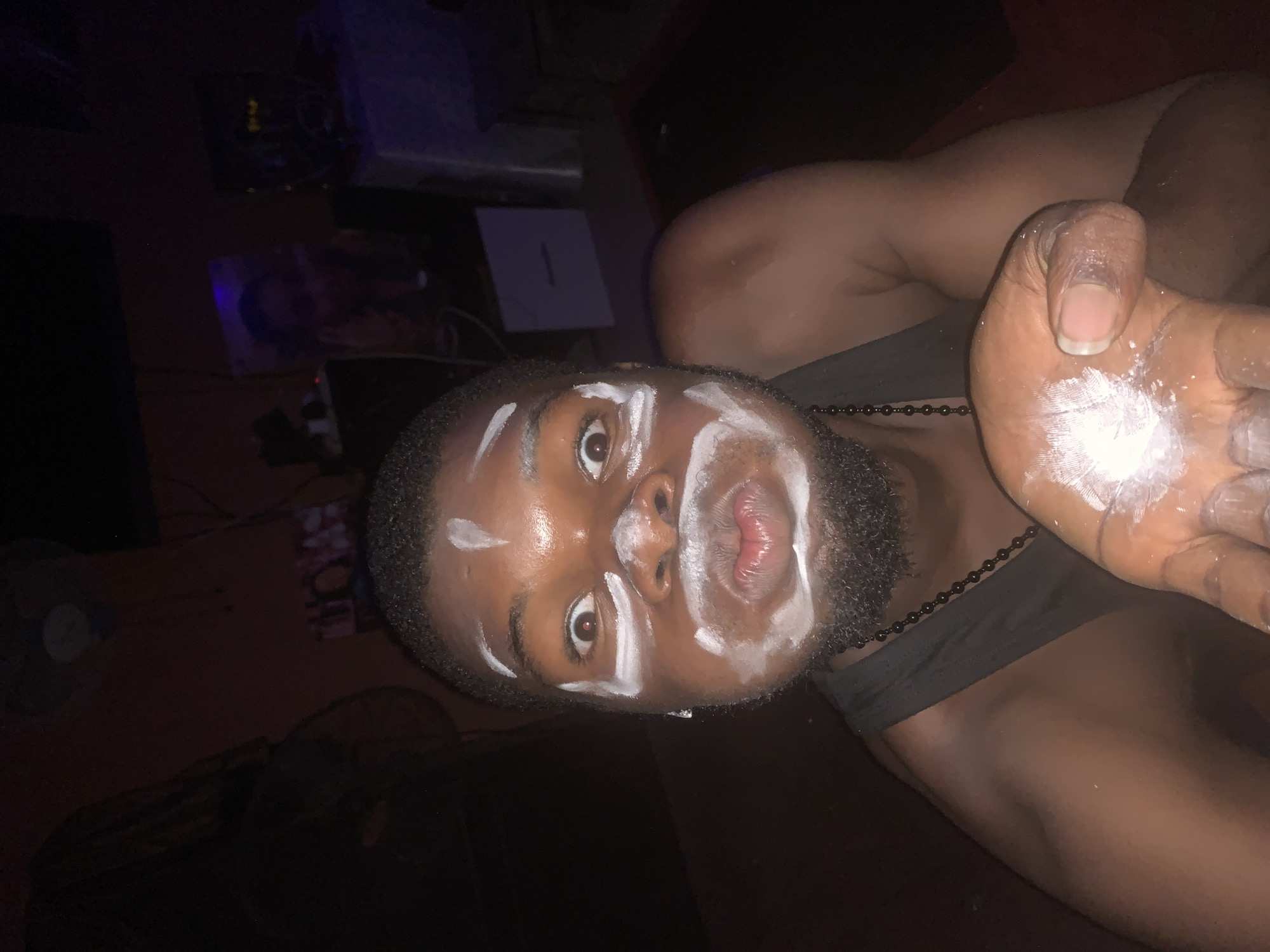 The image displays a young adult male with dark skin, situated indoors in a dimly lit setting, likely at night. He is positioned facing the camera with wide, direct eye contact and slightly pursed lips. His face is adorned with streaks of a white substance across his forehead, cheeks, chin, and upper lip, resembling face paint or a mask. His left hand, held up with the palm facing the camera, is also covered in the same white powder or substance. He wears a dark top and a dark beaded necklace around his neck.
The background is dark and largely out of focus, revealing hints of electronic devices or furniture. A white sheet or card with the number "1" is visible in the background, along with a partially visible fan blade on the left side of the frame. The overall expression on the subject's face appears playful or theatrical. This scene takes place in Onitsha, Nigeria.