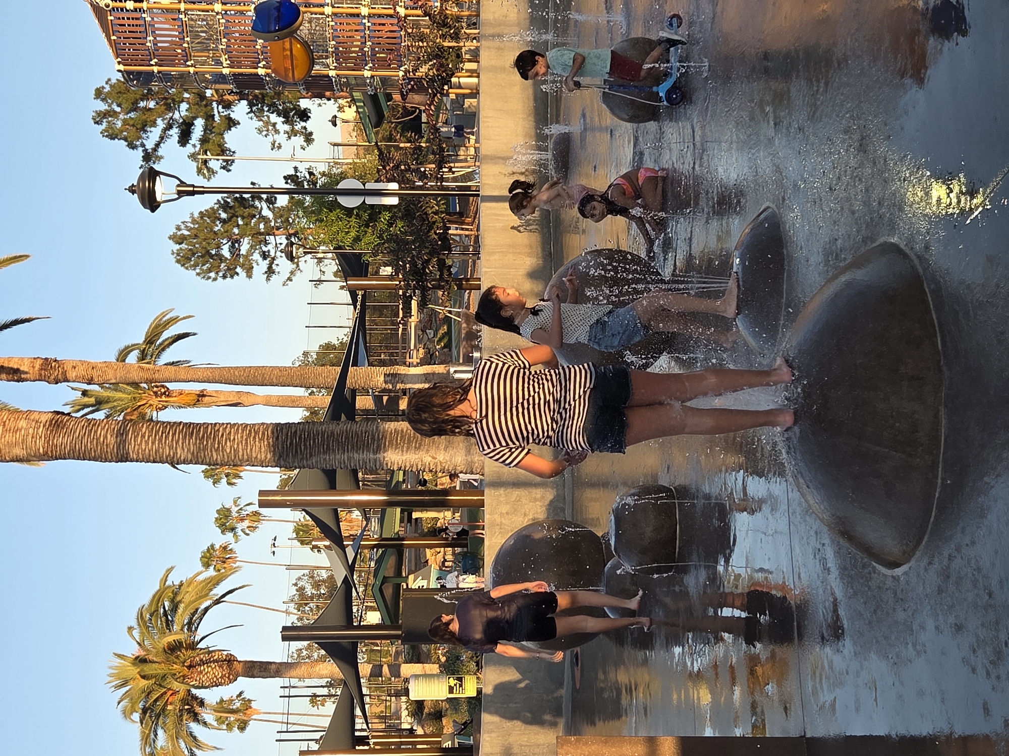 In Glendale, California, at a park's splash pad, several children are enjoying the water features on a sunny afternoon. The scene captures a moment of playful interaction as children run and play amidst the spraying water jets and large, dark, rounded stones that serve as interactive elements. In the foreground, a young girl in a striped shirt and denim shorts stands on one of the stones, facing away from the camera, with water splashing around her feet. To her left, another girl, also in a dark t-shirt and shorts, walks away, her reflection visible in the wet pavement. Other children are scattered throughout the splash pad, with one girl in a polka dot top and shorts reaching towards the water, another sitting in a pink swimsuit, and a young boy on a blue scooter riding through the spray. Palm trees and other greenery are visible in the background, along with elements of a playground structure. The lighting suggests it is late afternoon, casting long shadows.