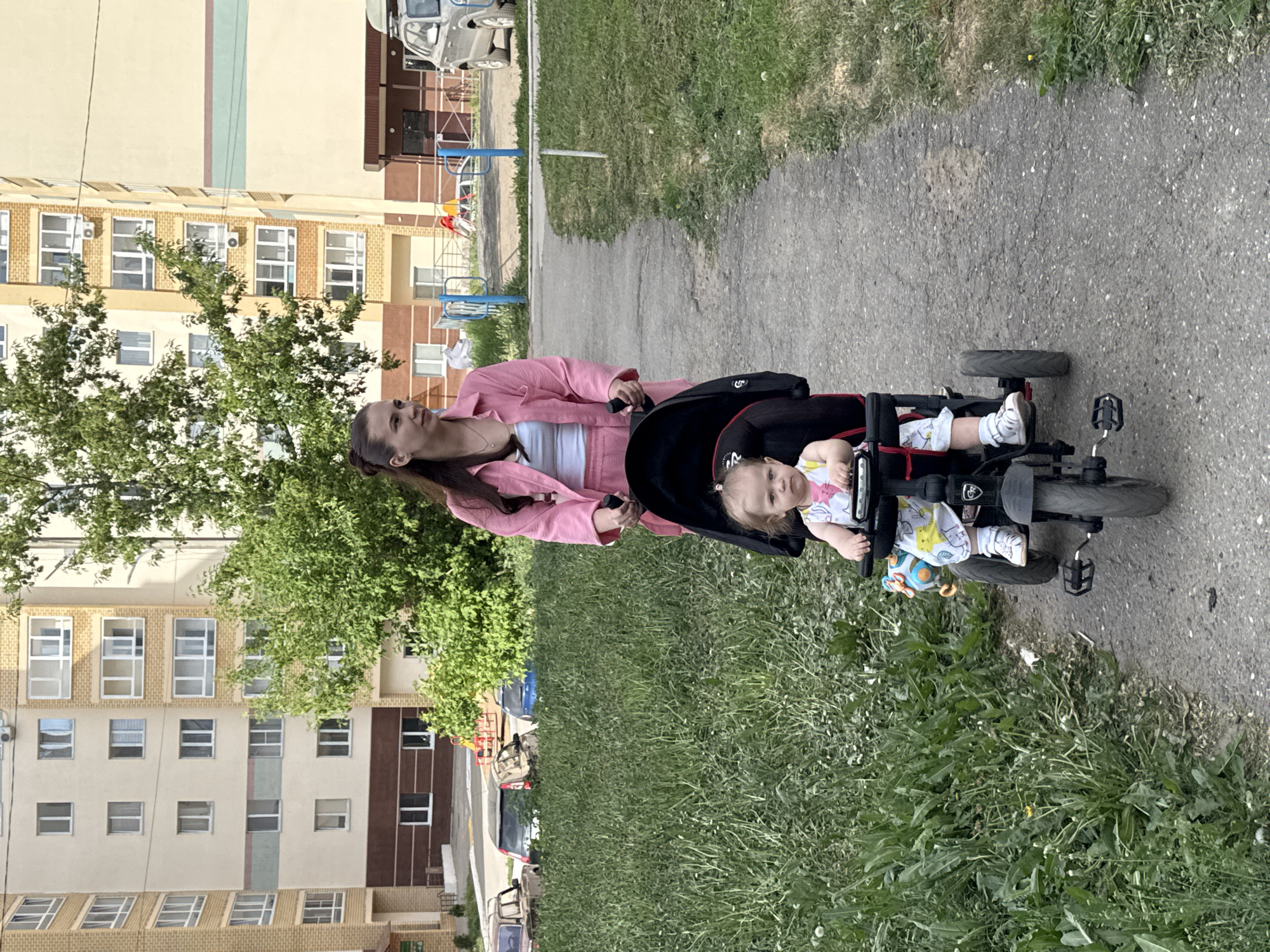 The image shows a woman pushing a child in a tricycle on a paved pathway. The woman is wearing a pink jacket and white top and has her hair pulled back. The child is seated in the tricycle, wearing a patterned white and yellow outfit, and looking at the camera.
In the background, a residential building with light-colored walls and multiple windows is visible. Trees line the pathway and lawn. Cars are parked nearby.
Based on the visual evidence, the location is likely a residential area with apartment buildings and green spaces in Chaikovsky, Russia.