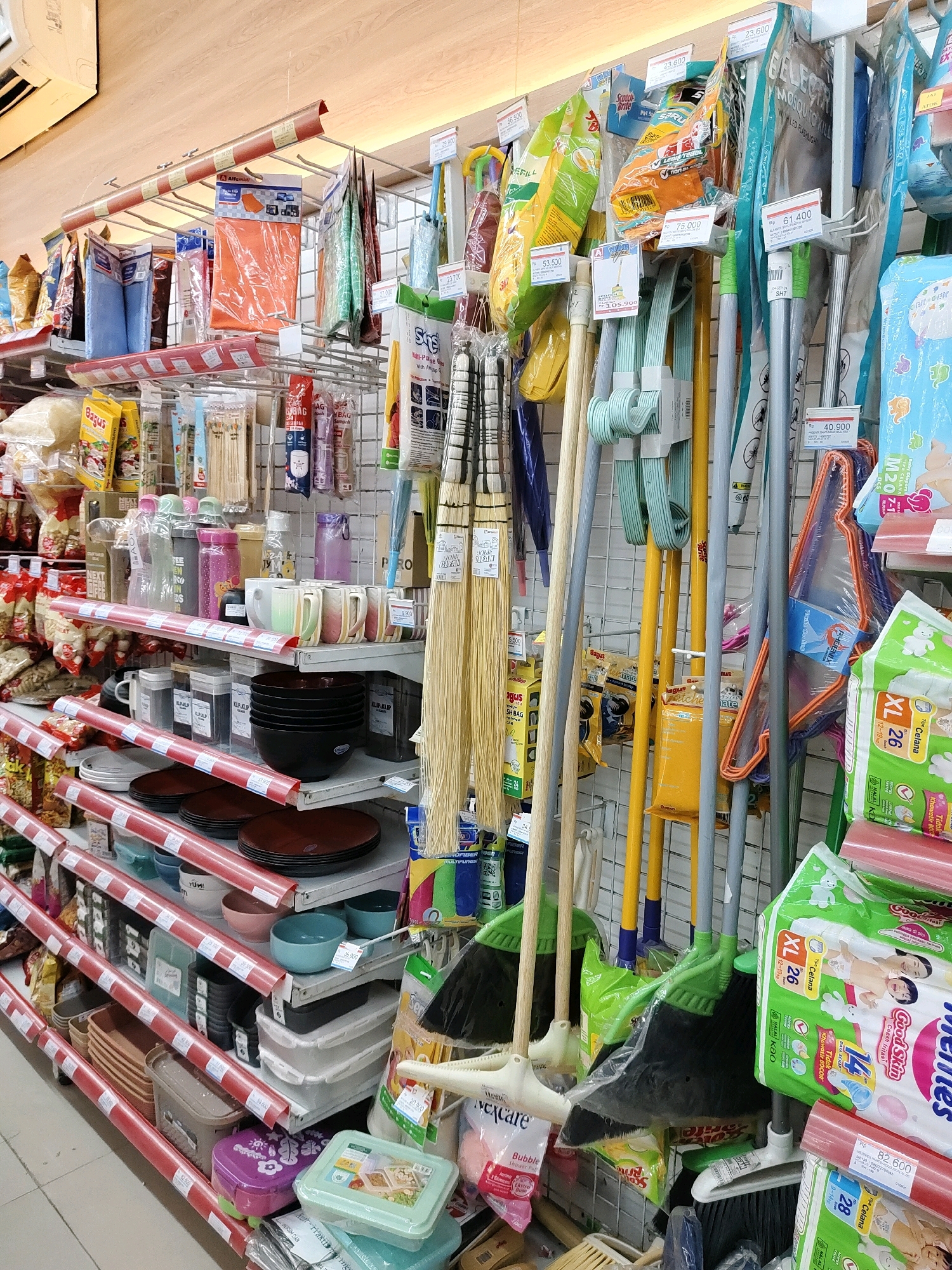 The image depicts a retail display in what appears to be a convenience store or supermarket, likely located in Probolinggo, Indonesia. The shelves are stocked with a variety of household items, including cleaning supplies such as brooms and mops, kitchenware like bowls and plates, food products in packaging, and personal care items like diapers. The items are arranged in an organized manner on white and red-trimmed shelves, against a backdrop of a light-colored wooden wall. There are no people visible in the image. The lighting suggests an indoor environment, typical of a retail establishment. The presence of Indonesian Rupiah (Rp) prices on some of the items further indicates the location.