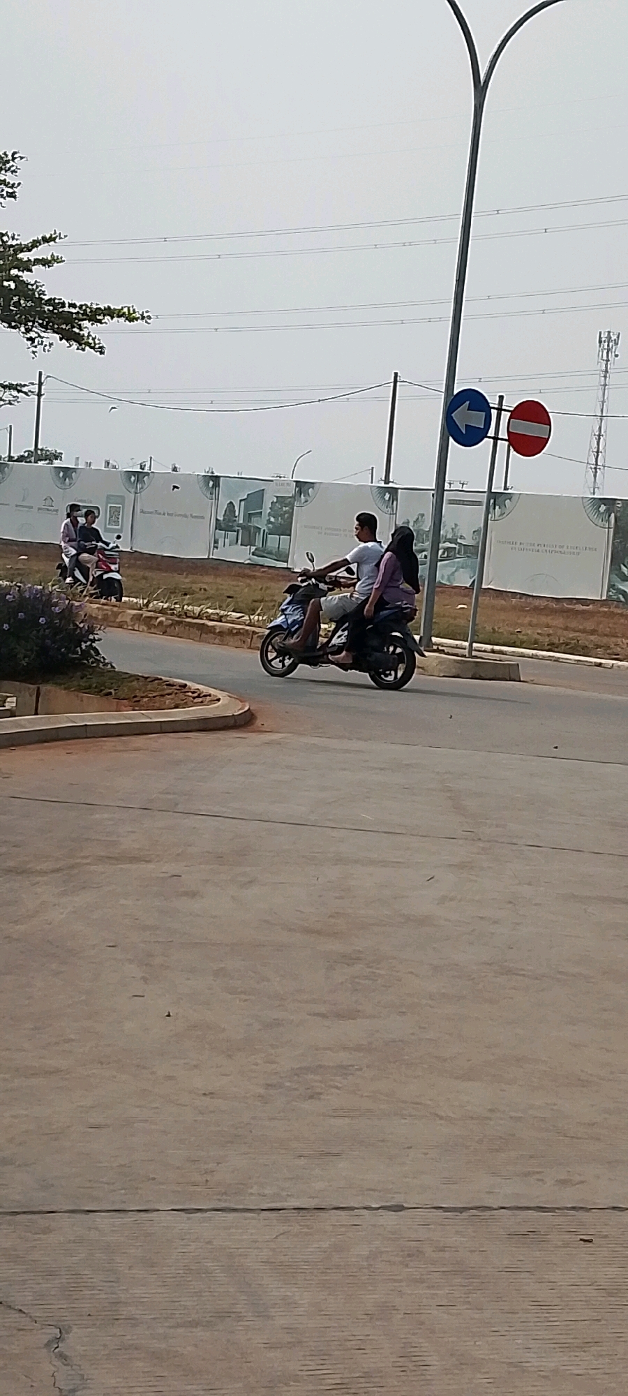 In Satriamekar, Indonesia, on an overcast day, two motorcycles are visible on a road. The foreground shows a concrete surface. In the middle ground, a motorcycle carries three people: a man driving, a woman behind him wearing a purple top and headscarf, and a second woman behind the first, also in a headscarf. To the left, a second motorcycle is also carrying two people, a driver and a passenger. They are riding on a paved road with a curb and grass verge. In the background, a large white banner with architectural renderings and text is erected. Power lines are visible stretching across the sky, and a cell tower is present in the distance. A street light with directional signs indicating left and no entry is positioned near the center of the image.