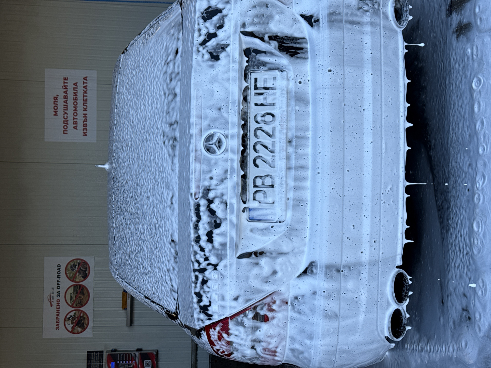 The image shows the rear of a car, heavily covered in white foam, likely undergoing a car wash. The car has a Bulgarian license plate that reads "PB 2228 HE". The make of the car appears to be Mercedes-Benz based on the central star emblem. In the background, there is a sign written in Bulgarian that instructs drivers to dry their cars outside the booth after washing. Adjacent to it is a sign that has a Bridge company's logo and "Забранено за off-road" (Forbidden for off-road), in Bulgarian with pictograms illustrating off-road vehicles that are prohibited. The car wash appears to be a self-service car wash facility. There is also a small kiosk with a screen visible in the lower left corner. The city of Sofia, Bulgaria is specified because the license plate is Bulgarian.