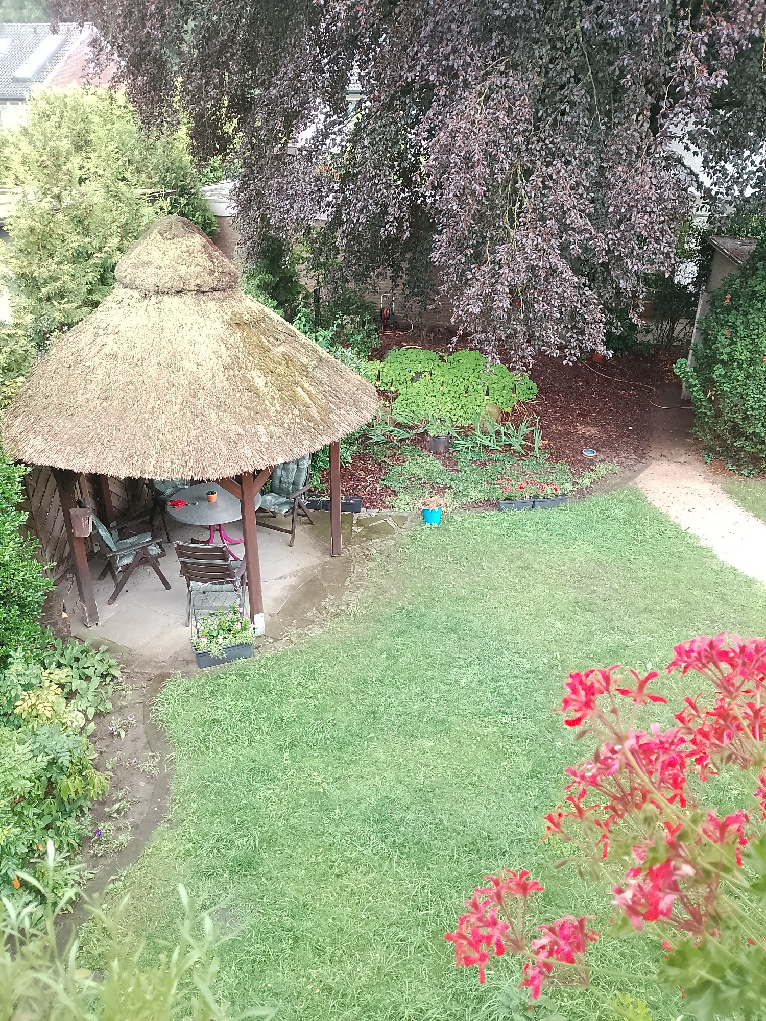 This image depicts a garden scene in Rheda-Wiedenbrück, Germany. In the center of the frame, a thatched-roof gazebo with a circular table and chairs is set on a stone patio. The gazebo is surrounded by lush green grass and various plants. A large tree with dark foliage overhangs the scene from the upper left. In the foreground, bright pink flowers are in bloom. The overall impression is a well-maintained, private garden space.