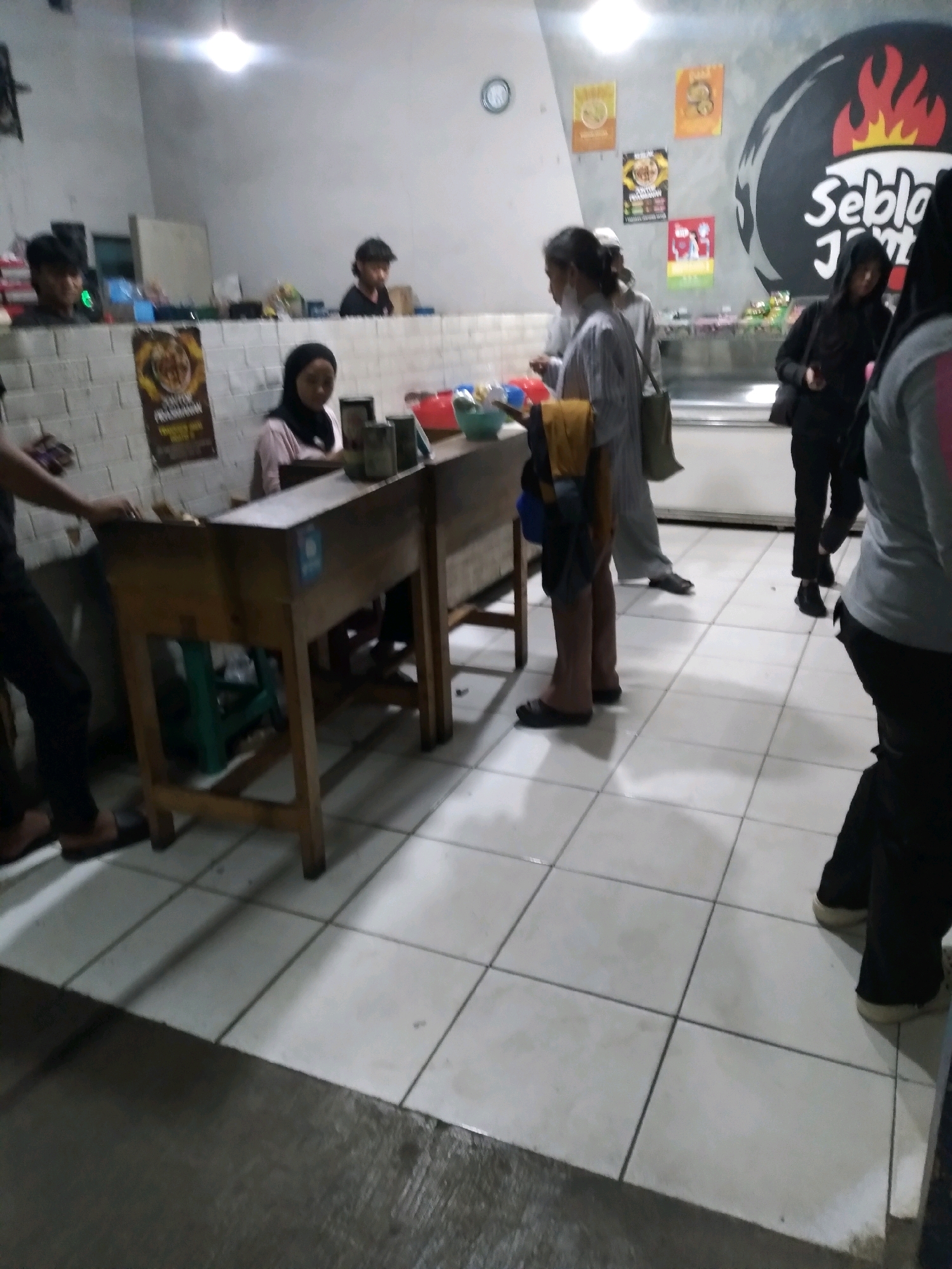 The image depicts a casual gathering or transaction taking place indoors, likely at a food stall or small eatery. The setting appears to be a commercial space with tiled flooring and a counter area. Several individuals are present, some interacting with the counter and others standing around. One person is seen at the counter, possibly making a purchase or being served. The lighting is somewhat dim, with a bright overhead light contributing to the illumination. The background shows a wall decorated with posters and a circular logo. The overall atmosphere suggests an informal, everyday scene.