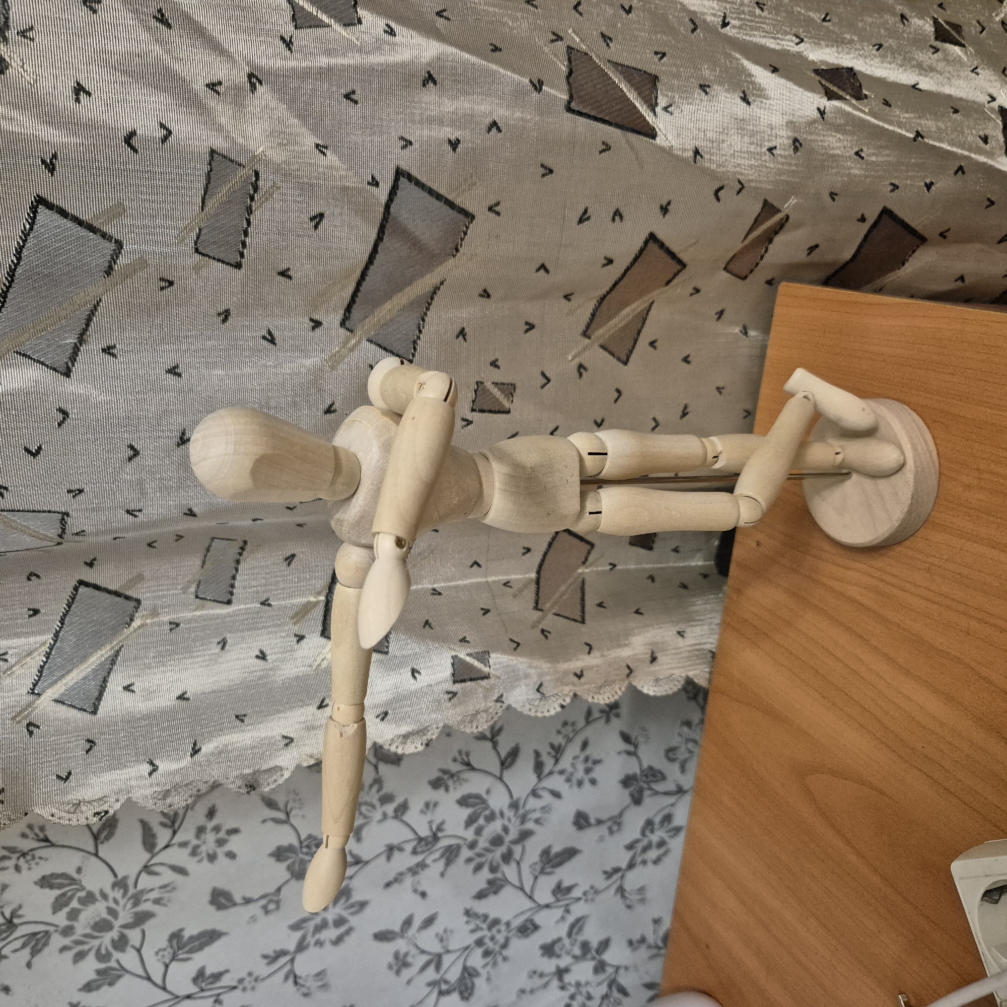 The image shows a wooden mannequin standing on a light-colored wooden surface. The mannequin is posed with its arms outstretched and bent at the elbows, as if in a dance or a gesture. It is positioned in front of a patterned curtain. The curtain has a floral pattern on the left side and a geometric pattern with rhombuses and small arrow-like shapes on the right side. The mannequin is the primary subject, and its joints are articulated. The background consists of the curtain and the wooden surface. There is no indication of the city of Liski, Russia, or any other specific geographical location.