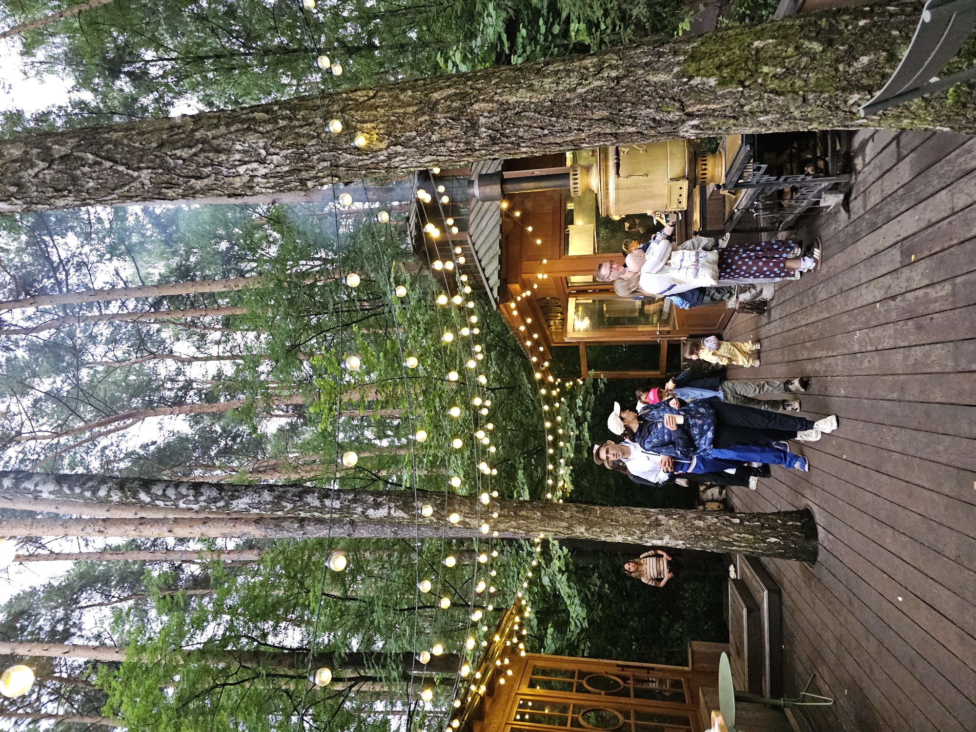 In Nemchinovo, Russia, a wooden walkway is set amidst a forest, illuminated by numerous string lights overhead. On the left, a wooden structure with large windows and glass doors appears to be a cafe or shop. A woman stands near the entrance. A group of people are walking on the walkway. One man is wearing a white jacket and blue pants, while a woman next to him wears a blue jacket and dark pants, holding a cup. A child in a pink hat walks beside them. Further down the walkway, a woman in a polka dot skirt holds a baby. Behind them, a large, golden samovar is situated on a stand next to a wooden kiosk or building, with smoke rising from its chimney. The scene is set in a dense forest with tall trees.