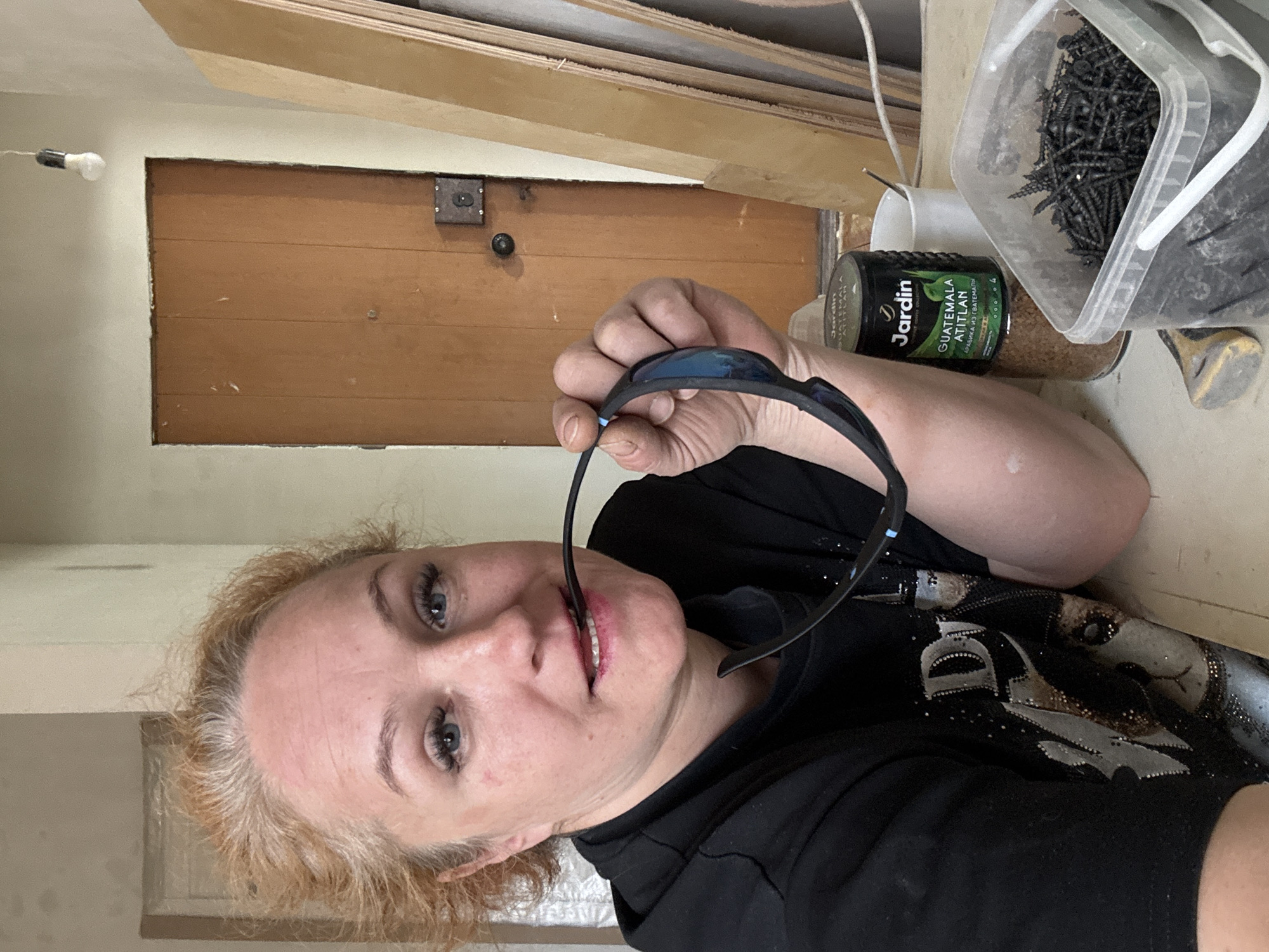 A woman takes a selfie indoors in Chaikovsky, Russia. She has light red/blonde, somewhat dishevelled hair and blue eyes, looking directly at the camera with a slight smile. Her lower lip shows a hint of red lipstick. She wears a black long-sleeved top with a sparkly, possibly embellished, design featuring a stylized letter 'D' and other shapes. She holds a pair of black sports sunglasses in her right hand, with one arm of the glasses near or in her mouth.
The setting appears to be a room under construction, renovation, or a workshop. To the left, a bare light bulb hangs from a wire against an off-white wall. Behind the woman, there's a wooden panel, possibly a door or partition, featuring a dark metal lock mechanism. On a surface to her right, various items are visible: a clear plastic container brimming with black screws, a black and green coffee can labeled "Jardin GUATEMALA ATITLAN" (with additional Cyrillic text below), and a glass jar containing a light brown granular substance, possibly sugar or sand. A small, light-colored tool or object is also present. Above, stacked wooden planks or plywood indicate a work or storage area. The overall impression is a casual moment during or after a period of work or activity.