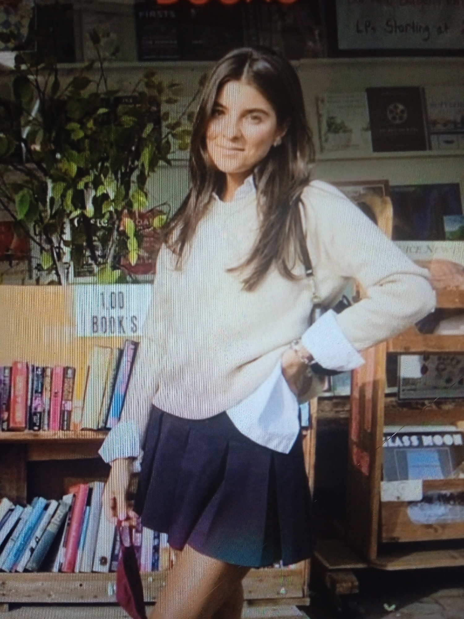 The photograph shows a young woman standing in a room with bookshelves. She is wearing a cream-colored sweater over a white collared shirt, and a dark pleated skirt. Her hair is dark brown and long, and she has a light complexion. She is smiling and looking towards the camera. The room appears to be a bookstore, with shelves filled with books visible behind and around her. There is also a plant on a shelf to her left. The lighting is somewhat dim, suggesting it might be indoors or during the evening. There is no indication of the location being in Chernivtsi, Ukraine.