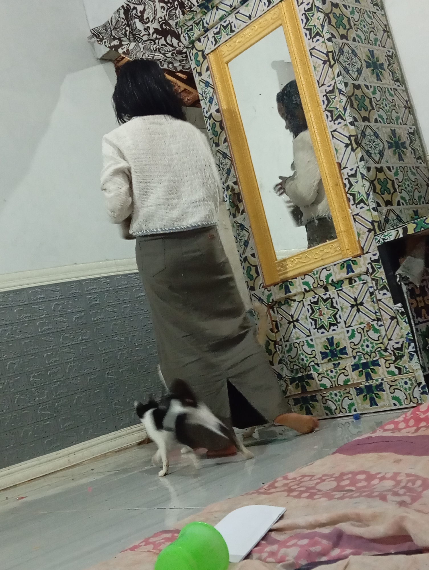 A woman, seen from the back, wears a white knit sweater and a long olive-green skirt. She stands near a wall with textured gray paneling, beside a decorative cabinet with a large gold-framed mirror. The cabinet is covered in colorful tiles featuring geometric patterns in green, blue, and white. A black and white cat is on the floor in front of the woman, appearing to move quickly. The floor is a light gray tile. In the background, a floral patterned fabric hangs from the ceiling.