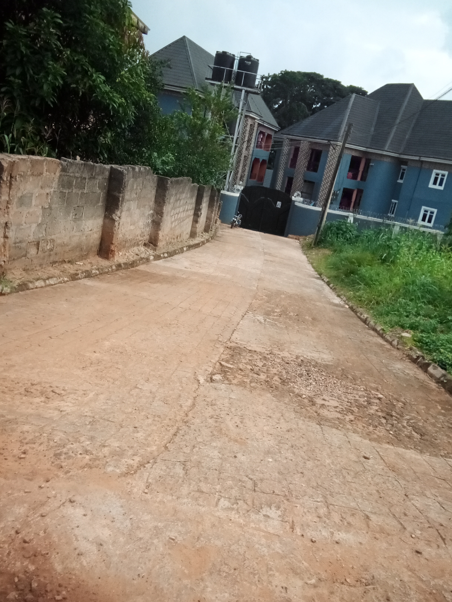 The image shows a street scene in Oko Town, Nigeria. A dusty, paved road leads away from the viewer, curving to the right. To the left of the road is a low, rough stone wall. Beyond the wall, dense green foliage is visible. Further up the road, on the right side, are two multi-story blue buildings with dark roofs. The buildings have multiple windows and balconies. In front of the buildings, a large black metal gate is visible, with a motorcycle parked to its left. Green grass and other vegetation grow along the side of the road, bordering the buildings. The sky is overcast.