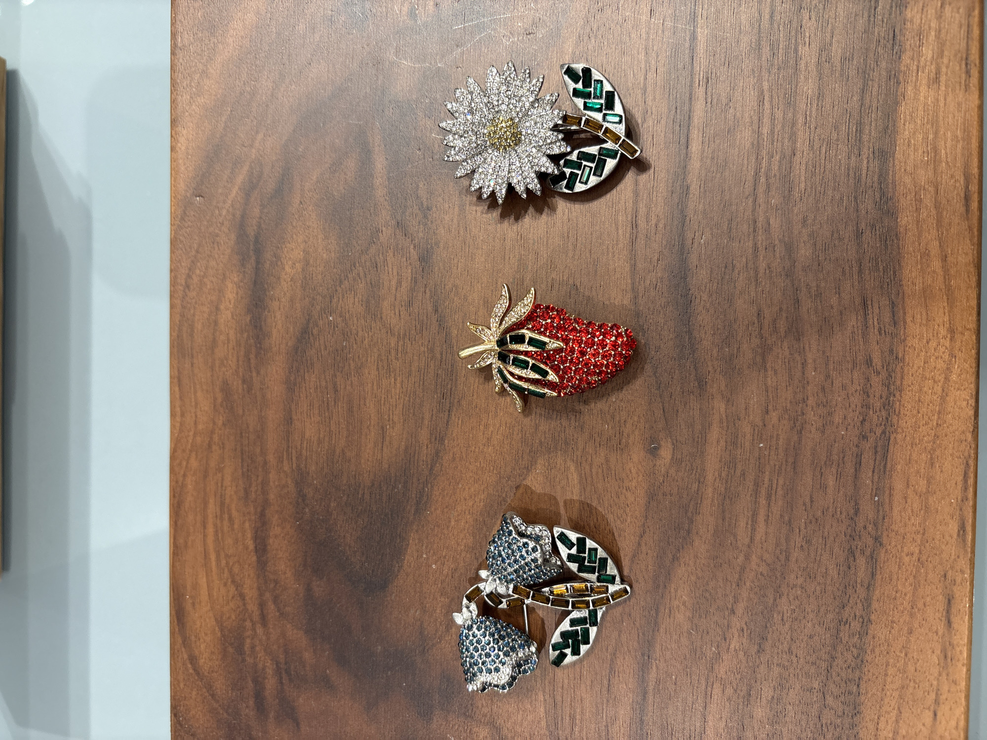 The image shows a wooden surface with three decorative brooches placed vertically in the center. From top to bottom, the brooches are:
1. A flower brooch with white/silver petals and a yellow center, accompanied by a separate leaf-shaped brooch with green and clear rectangular accents.
2. A strawberry brooch predominantly red with green accents, gold stem, and small clear gems on the stem and the body of the strawberry.
3. Two blue rose shaped brooches side by side with clear gems forming the flower, accompanied by a separate leaf-shaped brooch with green and clear rectangular accents.
The wooden surface is dark brown with a natural wood grain pattern. To the left of the wooden surface, a light gray surface is visible. There is no indication in the image that this is in Sofia, Bulgaria.