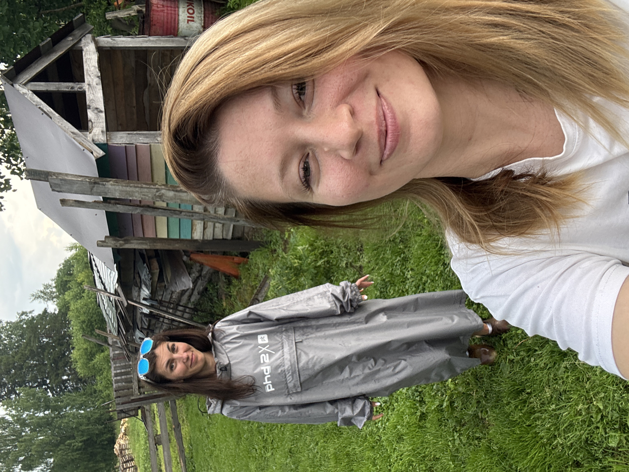 In a grassy, outdoor environment, likely in or near Вассята, Russia, two women are present. The woman in the foreground is facing the camera, her upper body visible, and she is smiling. She has blonde hair and is wearing a white t-shirt. Behind her, to the left, a second woman is visible from the shoulders down. She has dark hair and is wearing a long, grey hooded robe or poncho with "phd 2x pt" printed on the chest in white. She is also wearing brown shoes. In the background, there is a wooden structure with a corrugated metal roof, possibly a shed or small building, painted with horizontal stripes of various colors. A red barrel with white lettering that appears to be "OIL" is next to the structure. A wooden fence and trees are also visible in the background, suggesting a rural or semi-rural setting. The ground is covered in green grass.